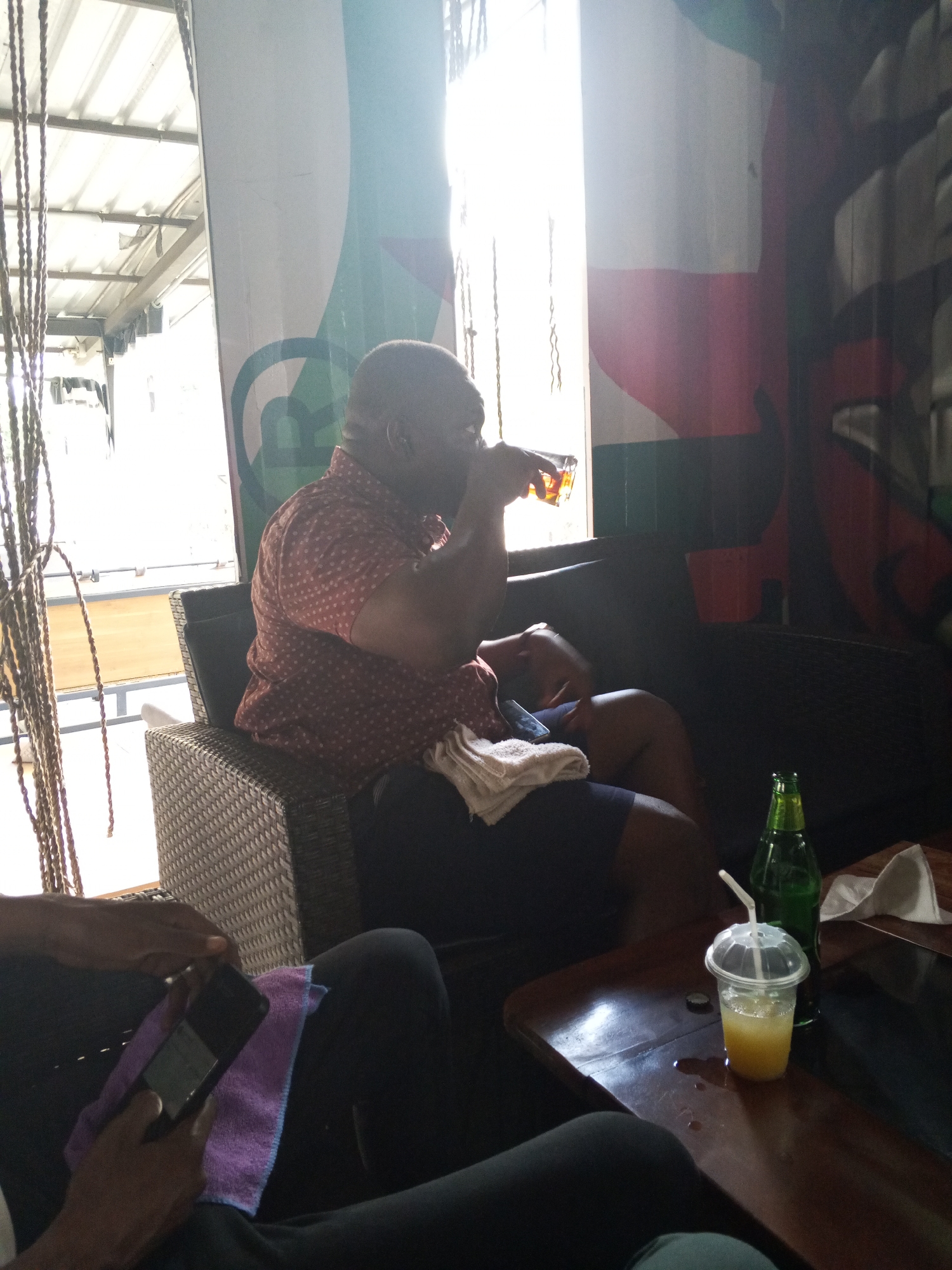 The image shows the interior of a lounge or bar, potentially in Abuja, Nigeria. An African man is sitting on a wicker chair with black cushions, drinking from a glass. He is wearing a dotted coral-colored shirt and dark shorts. A white towel rests on his lap.
A second individual is partially visible in the foreground, holding a smartphone. The phone is placed on a small, purple hand towel.
A dark, wooden table is in front of the subjects. On the table, there is a plastic cup with a straw, possibly containing juice, and a green glass bottle. Also on the table is a piece of white tissue or paper.
The background includes a decorative painting and hanging rope decorations. Light is streaming in from a window.