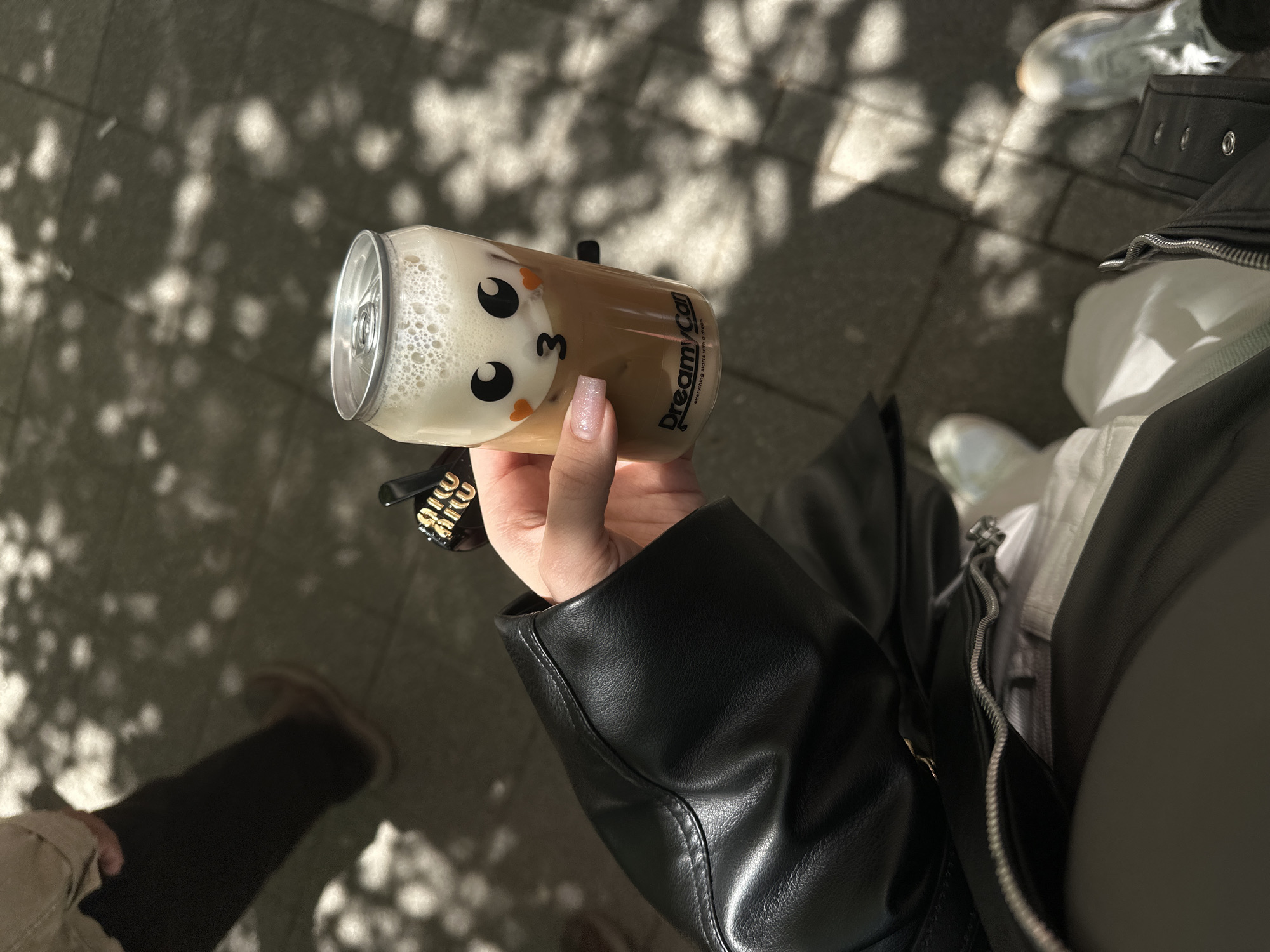 The image shows a person holding a "Dreamy Can" beverage in their hand, likely outdoors based on the background of dappled sunlight on a pavement. The can features a cartoon face design with foamy contents visible through the transparent part. The person is wearing a black leather-like jacket with a branded accessory attached to the sleeve or hand. Other individuals, partially visible, are wearing sneakers. The precise location cannot be determined without additional context. The city of Belgrade, Serbia, is not discernible from the visual information provided.
