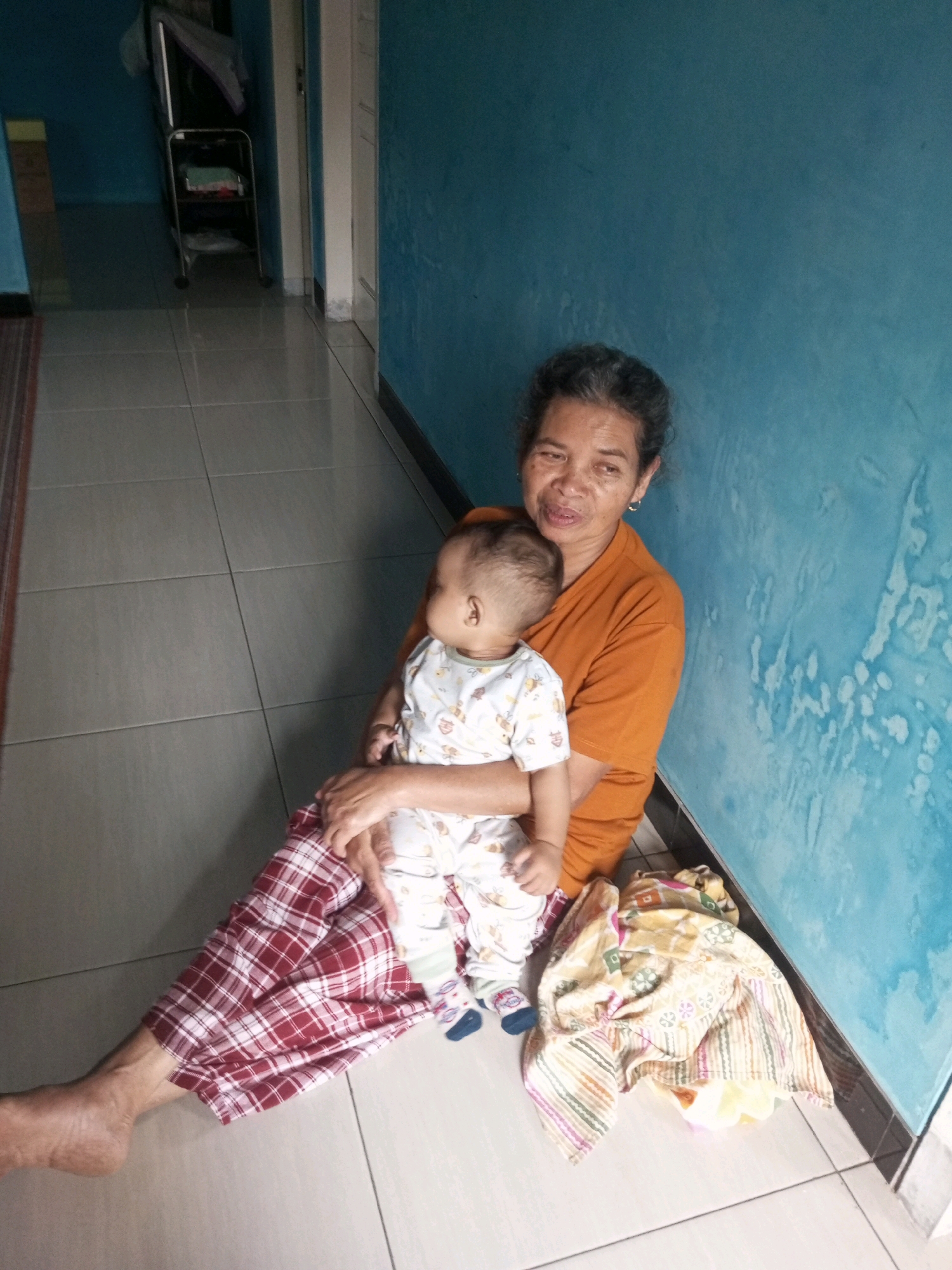 In Andongsili, Indonesia, a woman in an orange t-shirt and plaid pants sits on a tiled floor, holding a baby. The baby, dressed in a white onesie with a bear pattern, faces away from the camera. A colorful blanket lies beside them, and a metal shelf unit with items is visible in the background against a blue wall.
