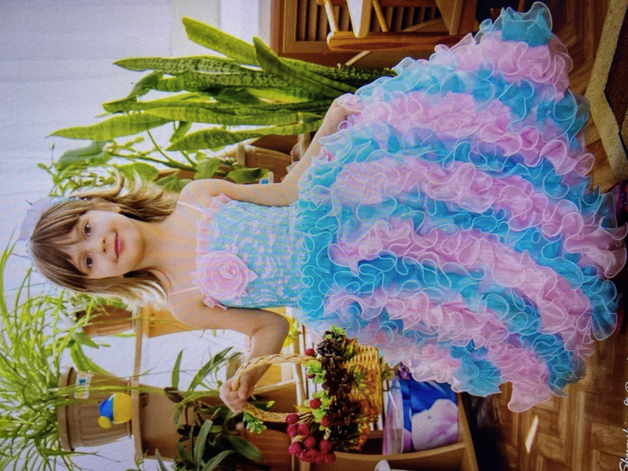 A young girl, appearing to be around five years old, stands indoors wearing a voluminous, layered dress in shades of pink and turquoise. The dress has a fitted bodice adorned with a pink flower. She holds a small woven basket filled with red berries and pinecones in her left hand. Behind her, several potted plants are visible, including a tall snake plant. The background consists of wooden shelves and a slatted wooden screen. The image's resolution suggests it might be a photograph taken with a low-quality camera or a screenshot from a video. There is no discernible connection to the city of Stavropol, Russia, based on the visual information provided in the image.