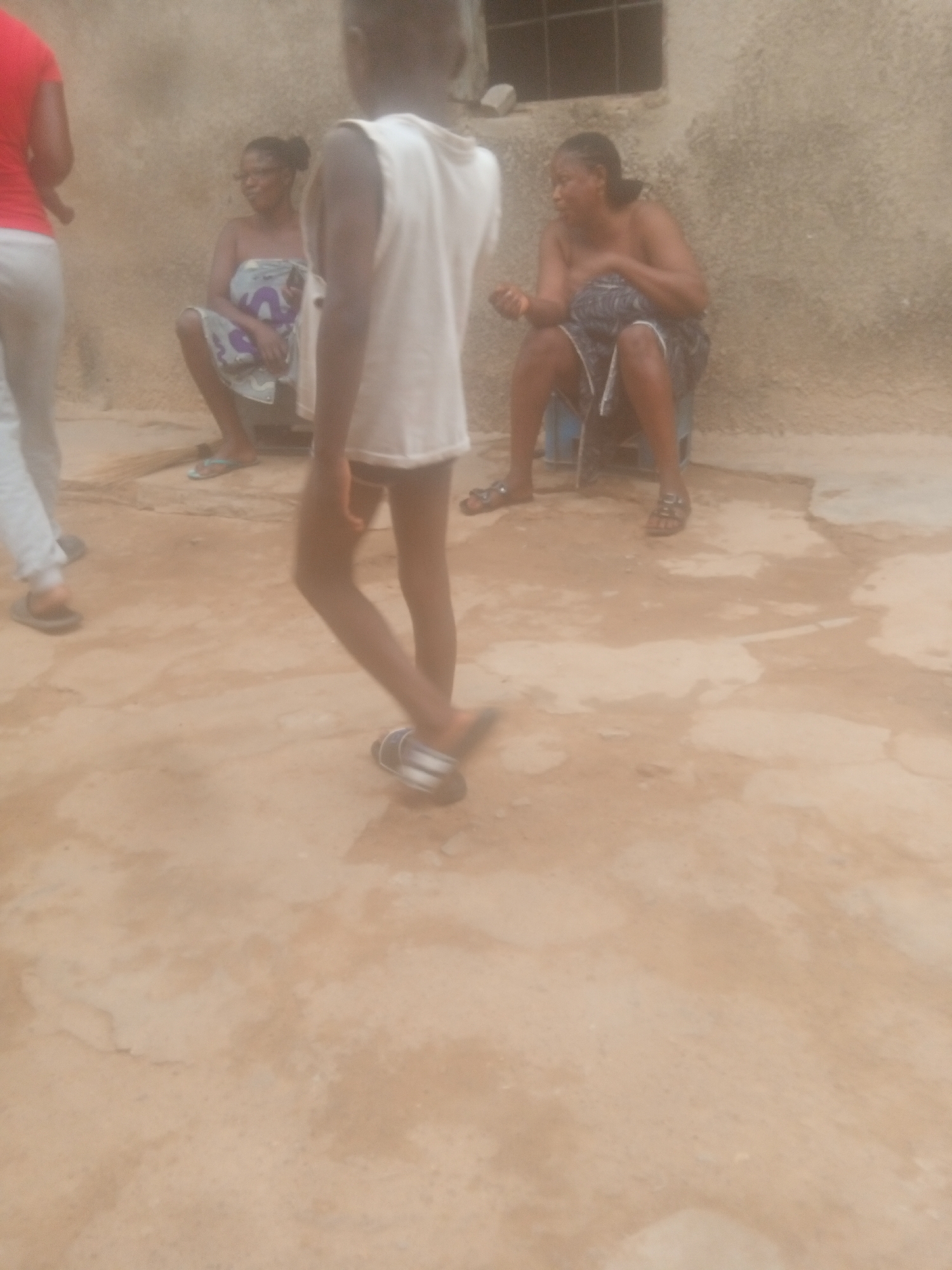 The scene depicts a somewhat overcast outdoor setting. The foreground is dominated by a tan, cracked concrete surface. A child wearing a white shirt and dark sandals is walking through the scene, partially obscuring the view of what is behind.
In the midground, there are two women sitting. One woman is wrapped in a patterned cloth and the other is seated on a blue stool, also with a patterned cloth wrapped around her.
Behind the women is a faded, textured wall, and a small barred window can be seen near the top of the image. The location appears to be in a city of Unknown, Unknown.