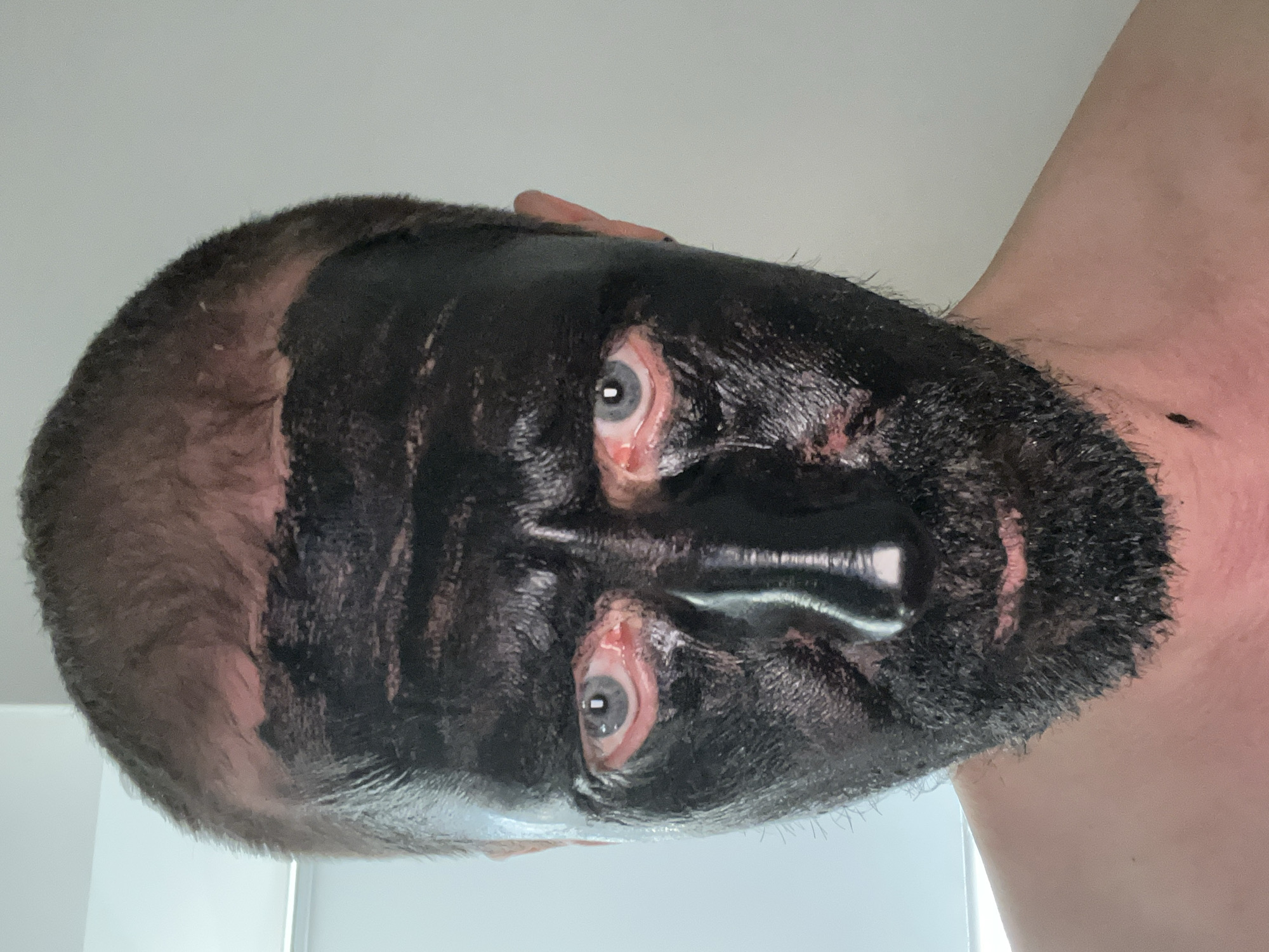 The image is a close-up selfie of a man's face, from the shoulders up. He has short, dark hair and is wearing a thick, black substance that covers most of his face, including his forehead, cheeks, nose, and chin. His eyes are visible and appear to be blue, with some redness around them. The black substance is applied unevenly, with some areas appearing thicker than others. His mouth is partially visible beneath the black substance, and he has a beard that is also partially covered. The background is a plain, light-colored wall. There is no discernible location in the city of Kyiv, Ukraine.
