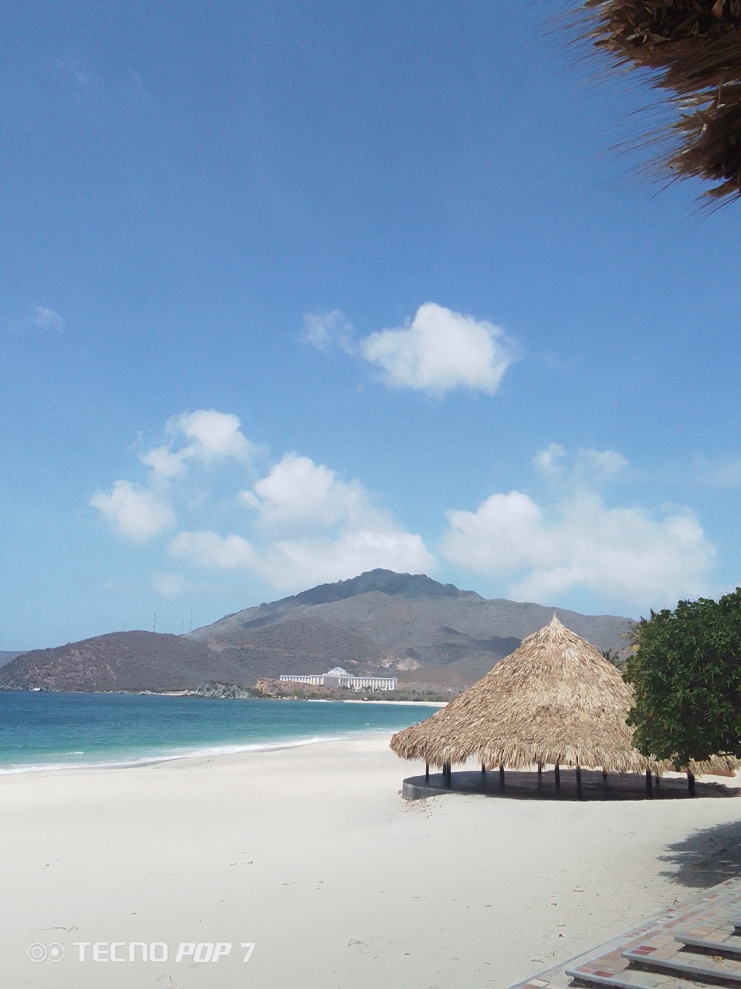 The image captures a scenic view of a beach in Pedro González, Venezuela. The foreground features a sandy beach with a thatched-roof structure providing shade. In the background, a mountain range rises against a clear blue sky with scattered clouds. A building complex can be observed along the coastline in the distance. The turquoise water of the sea contrasts with the white sand of the beach. The shot is taken in daylight, capturing the natural beauty of the coastal landscape.
