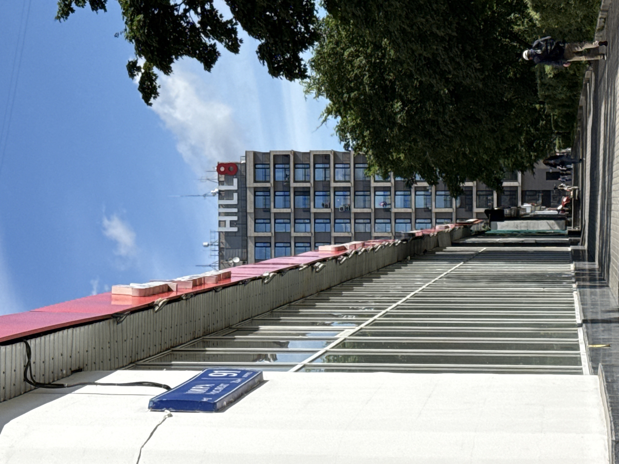 The image shows a street scene in Moscow, Russia, on a sunny day. In the foreground, on the left, is the side of a white building with a red overhang. A blue street sign with Cyrillic lettering and the number "91" is attached to the white wall. The building has a long row of large windows with vertical framing.
In the middle ground, a modern, multi-story residential building stands with a sign at the top that reads "HILL 8" in white letters against a gray background, with a red circle containing the number "8" next to the word "HILL". The building has a grid of many windows, some of which appear to be balconies with blue railings. To the right of this building, lush green trees form a dense canopy, obscuring part of the sky.
In the background, more trees and sky are visible. On the right side of the street, a man wearing a backpack and a white cap is walking away from the camera. Further down the street, several other people are walking. The street appears to be paved with cobblestones or similar material, and there are visible shadows cast by the sunlight. The overall impression is a contemporary urban environment with a mix of architecture and nature.