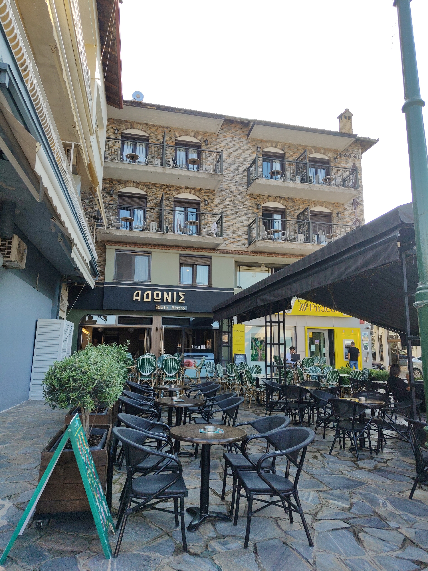 This is a daytime, outdoor, medium-shot photo taken on a paved street in Litochoro, Greece, with a building that houses a cafe and what appears to be a bank in the background. The building facade is made of stone and has multiple balconies with railings. The cafe, named "ΑΘΩΝΙΣ Cafe Bistro," has an awning covering part of its outdoor seating area. Many tables and chairs are set up outside the cafe, some of which are occupied by people. The chairs are black and wicker, and the tables are round with dark tops. A prominent green A-frame sign stands in the foreground, partially obscured by a potted tree.
In the background, a bright yellow building with a prominent Piraeus Bank logo is visible. The sky is bright, suggesting it is a clear day, likely midday. A few people are visible in the scene: one person sits at a table with a laptop outside the cafe, another person is standing near the ATM outside the bank, and a few other patrons are seated at tables under the awning. The overall atmosphere seems relaxed, with people enjoying the outdoor seating area of the cafe. The paving stones on the ground are large and irregular in shape. A green lamppost is visible on the far right of the frame.