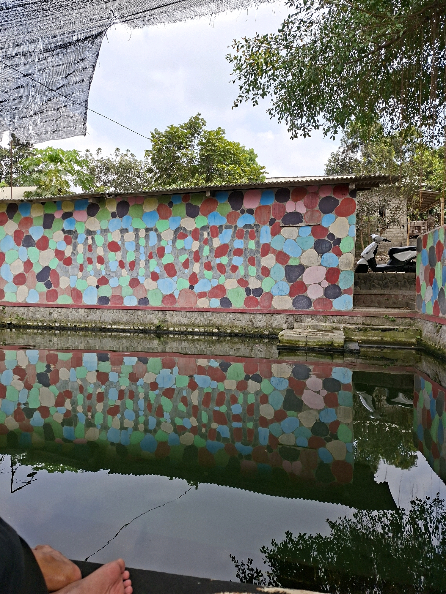 The image depicts an outdoor scene featuring a colorful, pebble-patterned wall that runs along the edge of a dark body of water. The wall is adorned with irregularly shaped, painted stones in various colors, including red, blue, pink, green, and black. Below the wall, the water reflects the vibrant pattern, creating a mirror image of the wall's design.
In the background, there is a glimpse of a building with a corrugated metal roof. To the right of the building, a white scooter is parked on a set of stairs leading up to the structure. Trees with green foliage are visible behind the wall and above the building, suggesting a natural or semi-natural environment.
A black mesh shade structure is stretched across the top of the image, casting shadows and partially obscuring the sky, which appears to be partly cloudy. In the foreground, a person's bare feet and legs are visible, resting by the edge of the water. The overall impression is that of a recreational area, possibly a park or a garden feature, in Blitar, Indonesia.