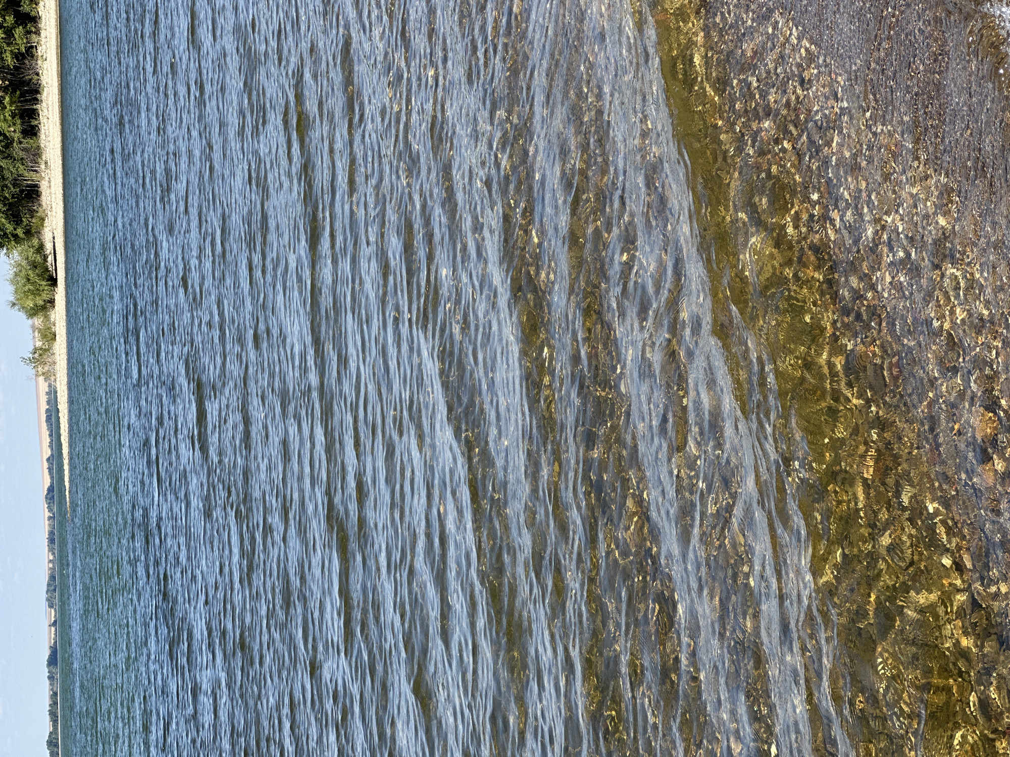 The image depicts a body of water, likely a lake or reservoir, with a visible shoreline in the foreground. The water is clear enough to see the rocky bottom near the edge. Gentle waves create ripples on the surface. In the background, a distant shoreline with trees and a field under a partly cloudy sky is visible. The scene appears to be a natural landscape, possibly in a rural or park-like setting. No people or specific structures are identifiable in the image.