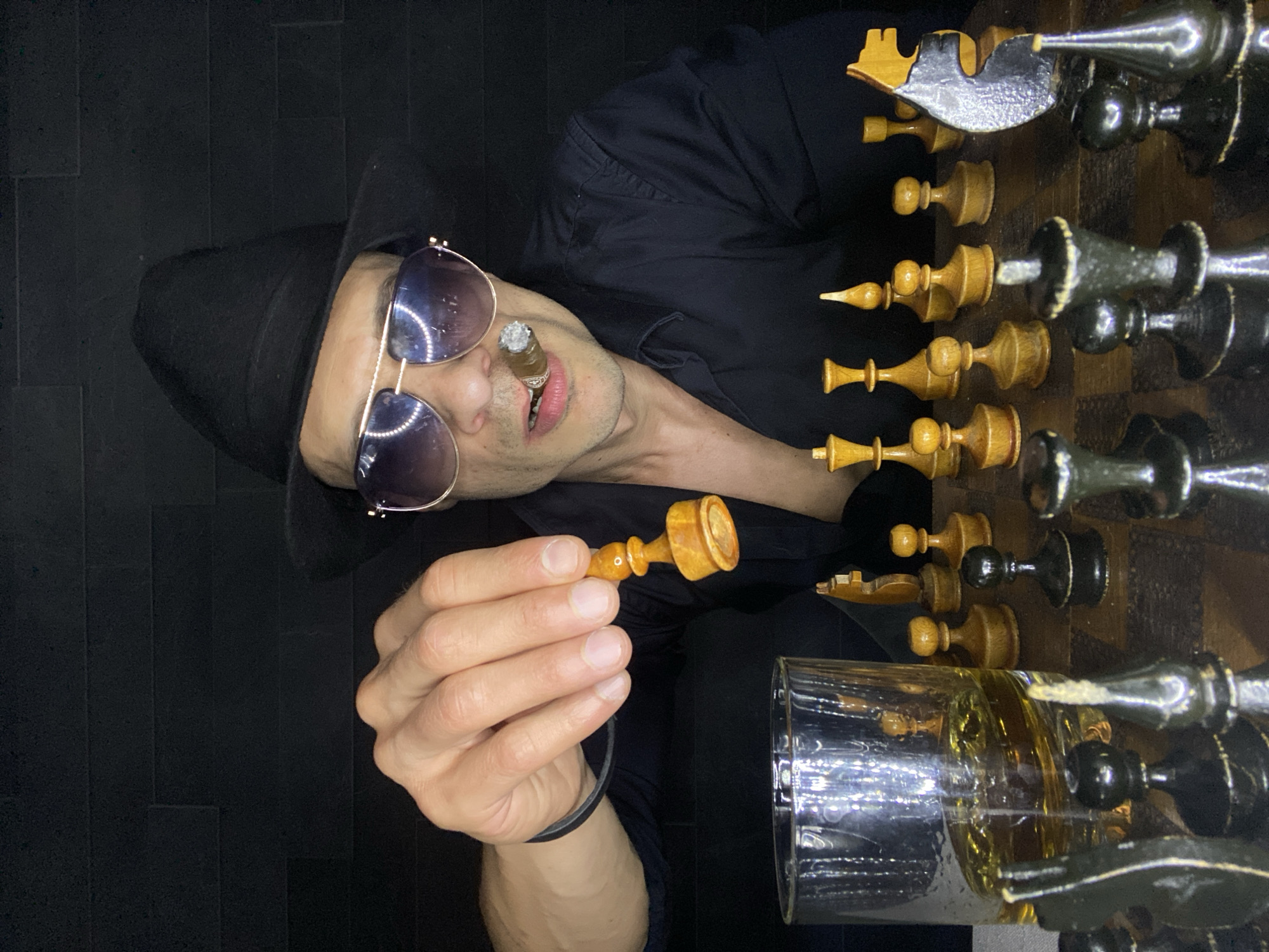 A person wearing a black hat and sunglasses is holding a chess piece and smoking a cigar while standing next to a chess board and a glass of amber liquid. The background is black. The location is not discernible based on the image.