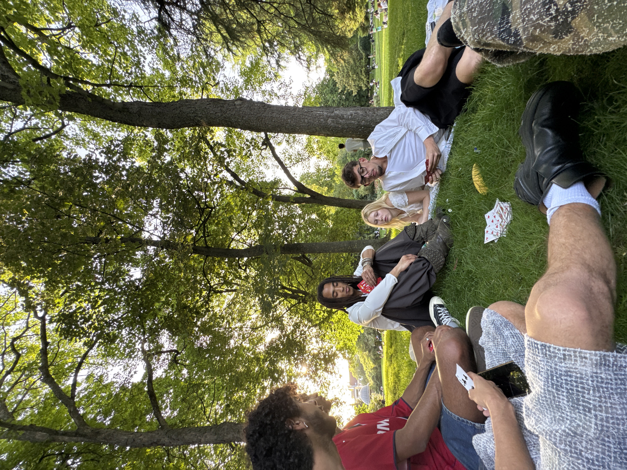 A group of people is sitting on the grass under trees. A man wearing a white shirt and glasses is lying on a white sheet, with a blonde woman next to him. Another person with long dreadlocks is sitting with their legs crossed. A person wearing a red shirt is looking at the camera. Another person is holding a phone. Playing cards are scattered on the grass. The location is not identifiable as Paris, France from the visual information available.