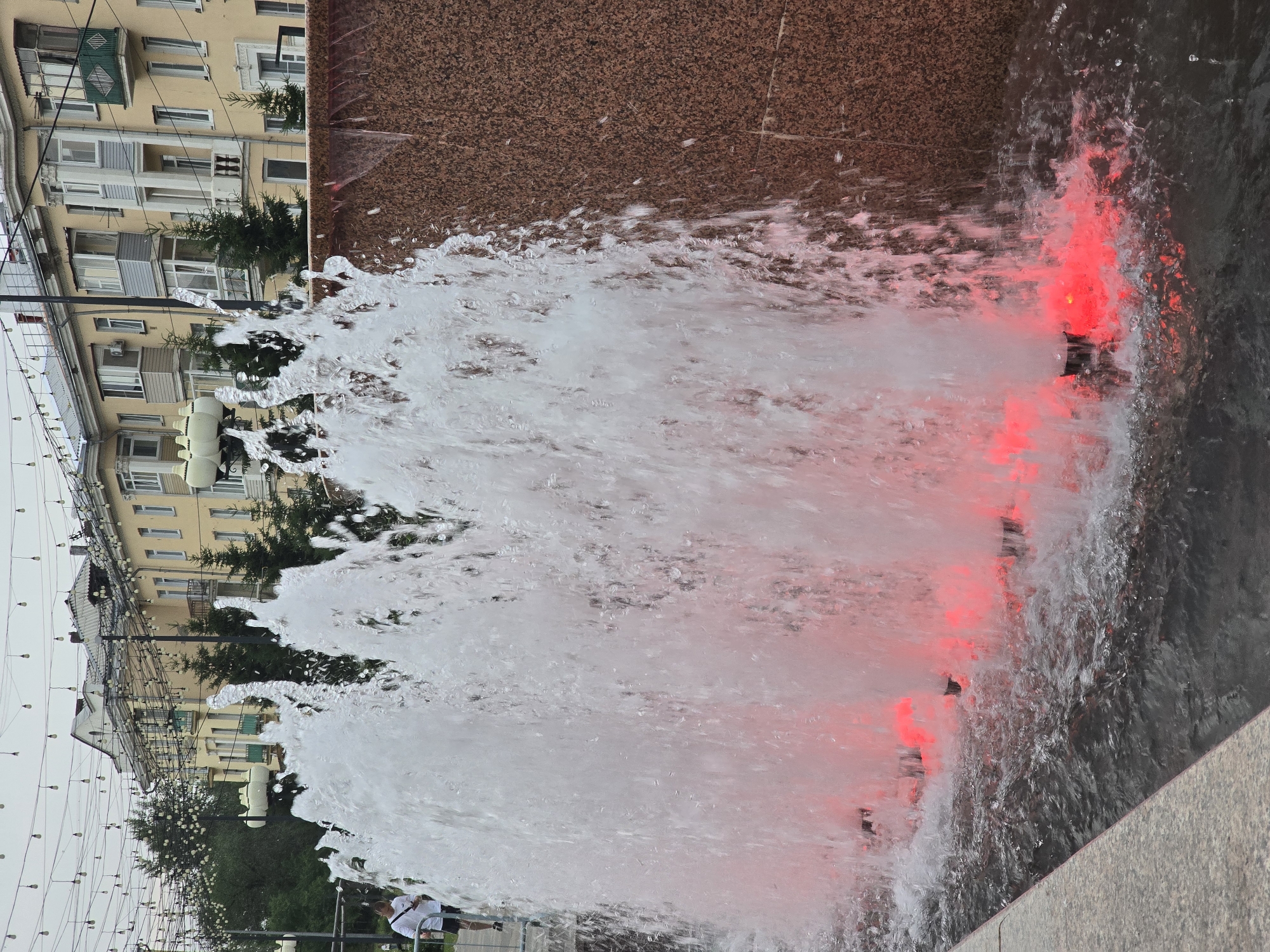 In the foreground of the image, a fountain sprays water upwards, illuminated from below by red lights, creating a vibrant visual effect. The fountain is positioned in front of a textured, reddish-brown wall. In the background, several multistory buildings with balconies and windows are visible, suggesting an urban setting. Overhead, strings of lights are suspended between poles, indicating a decorative or festive lighting arrangement. A person wearing a white t-shirt and dark shorts is visible on the left side of the image, walking along a paved area. The sky is overcast.