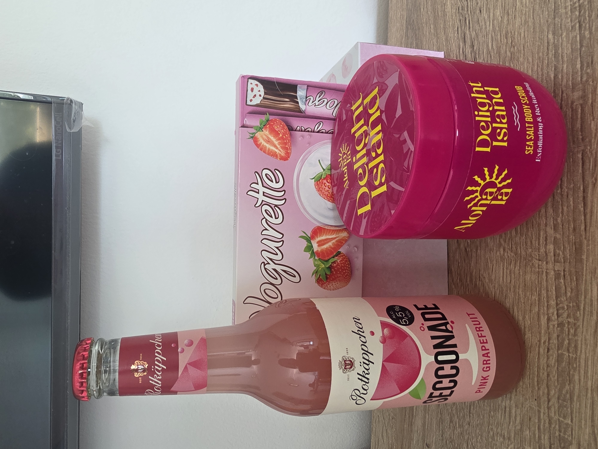 The image displays a still life arrangement of pink-hued products on a wooden surface. In the foreground, a tall glass bottle of "Rotkäppchen Secckonade Pink Grapefruit" stands to the left, its label featuring a stylized wine glass and the brand's name. To the right, a bright pink plastic tub of "Aloha la la Delight Island Sea Salt Body Scrub" is visible. Behind these, a pink cardboard box labeled "Voguette" with images of strawberries and what appears to be chocolate bars is positioned. The background shows a white wall and a portion of a dark screen with "LG NanoCell" branding visible at the bottom edge. There are no people present, and no identifiable landmarks or specific urban features suggest a location in Munich, Germany. The lighting appears to be natural, coming from the left.