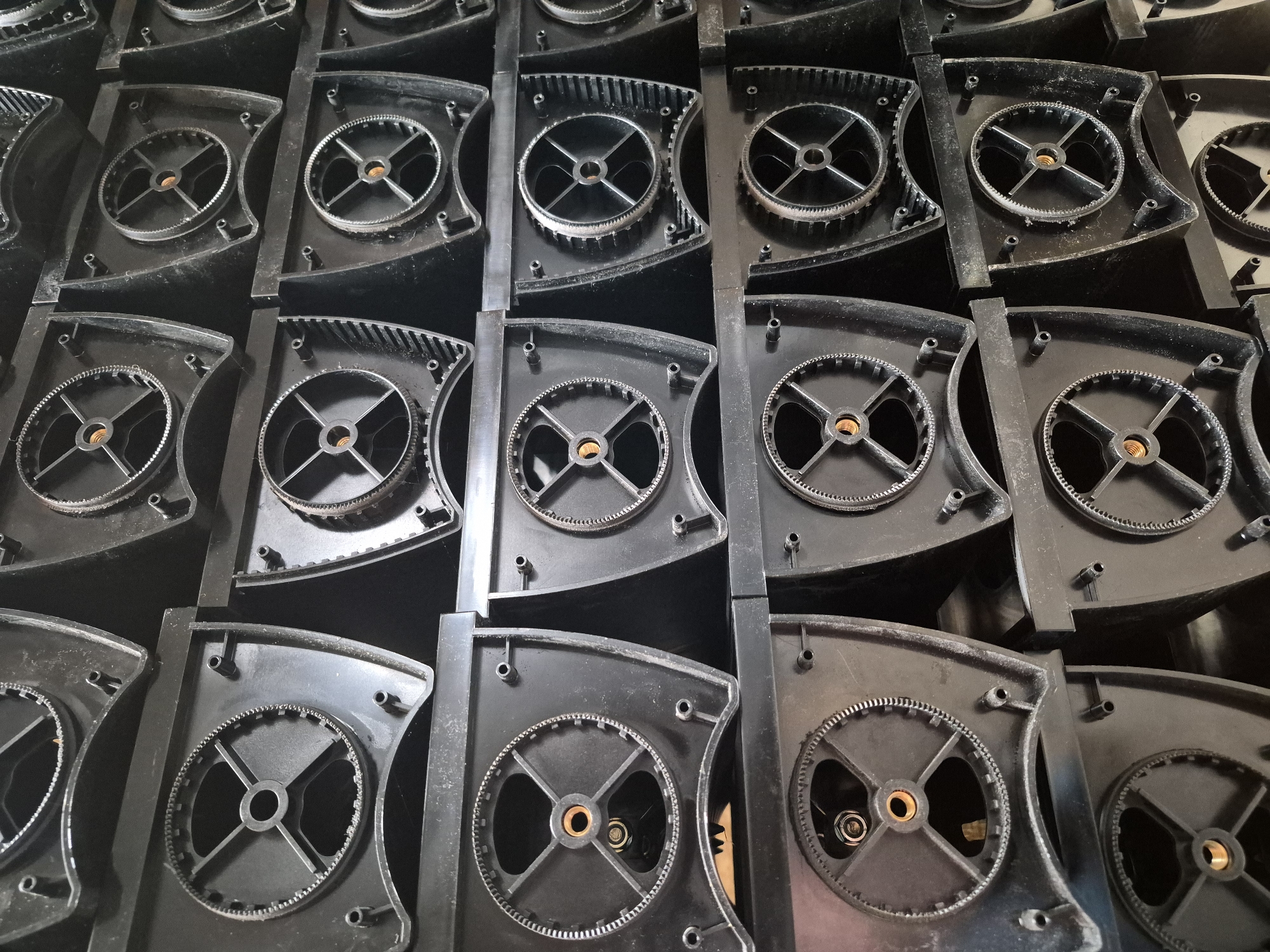 The image shows a stack of black plastic components, likely parts of an industrial machine or appliance. Each component features a circular gear mechanism with spokes radiating from a central hub. These components are arranged in a grid-like fashion, with several layers visible. The lighting is somewhat dim, and the overall impression is one of industrial storage or manufacturing. There are no people or animals in the image. The location within the city of Masów, Poland, is not discernible from the visual information provided in the image.