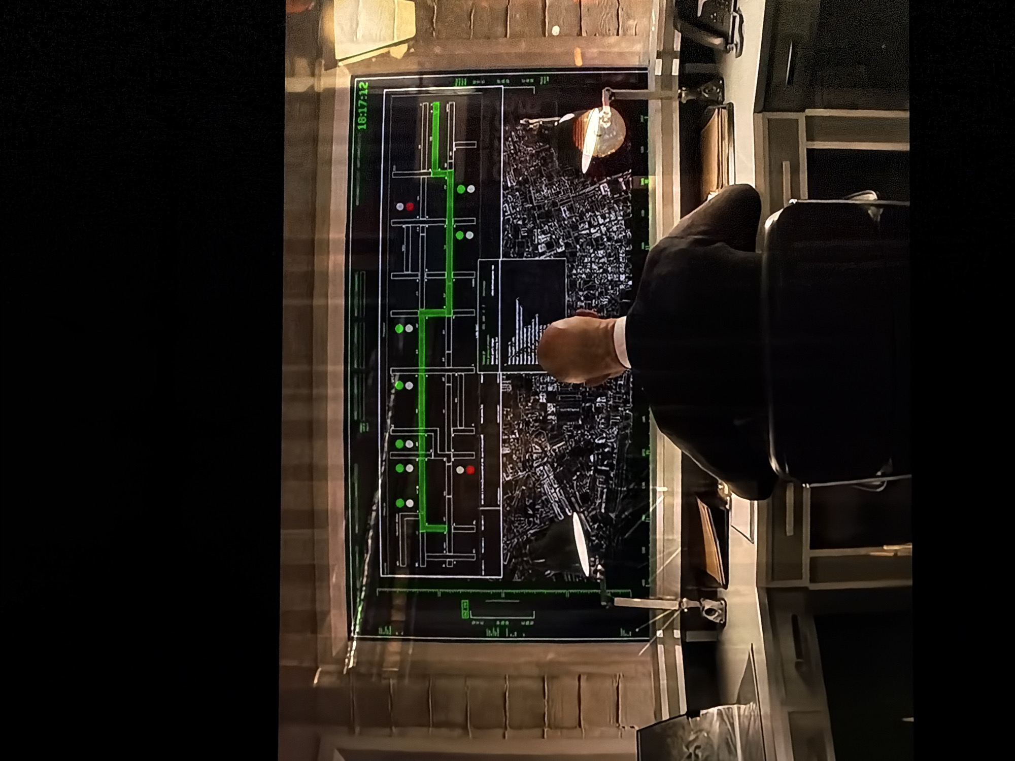 The image displays an overhead view of a person seated at a large, illuminated desk in a dimly lit indoor environment, possibly an office or a control room. The individual, seen from behind, is wearing a dark suit jacket and appears to be observing or interacting with a prominent, large digital display embedded in the desk surface.
The display features a dark background with bright green and white/light gray graphical elements. On the left side, a detailed floor plan or architectural layout is shown, marked with a significant green path and several circular indicators in both green and red, suggesting tracking or status points. The right side of the display presents an aerial or map view of an urban area, depicted in a monochromatic style with intricate street patterns and building outlines. Below these sections, a bar graph or similar data visualization is visible.
A desk lamp with a metallic shade provides a focused light source, reflecting off the glossy surface of the display and the desk. The person's right hand is positioned near the lower right portion of the screen, suggesting interaction with the interface.
A notable detail is the timestamp "18:17:12" displayed in green text at the top-left corner of the screen. Other smaller, illegible text and symbols are scattered across the various sections of the interface. The overall atmosphere conveys a sense of focused work or strategic analysis. The location context provided is Sofiivska Borshchahivka, Ukraine.