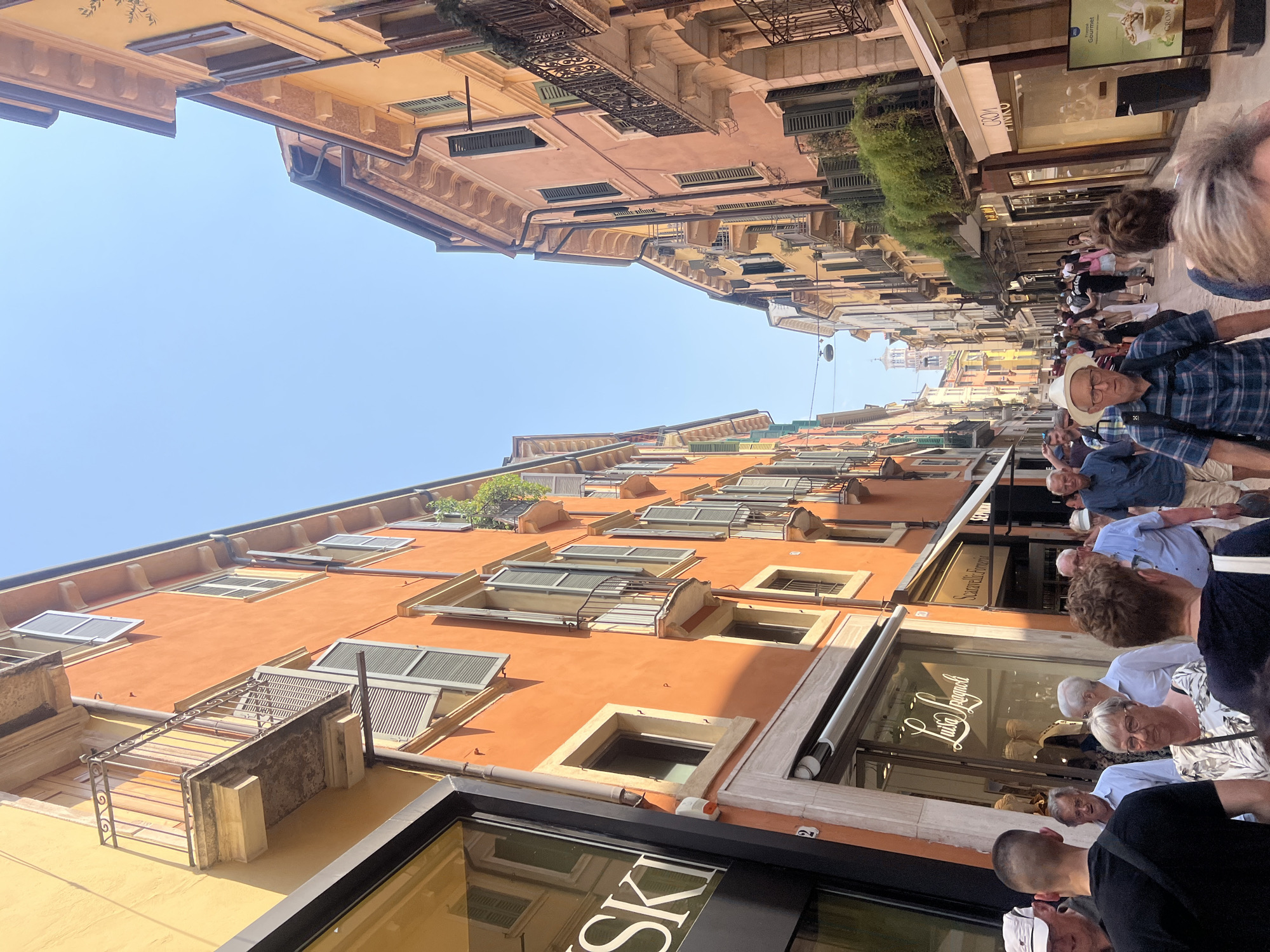 The image captures a street scene in an unidentified city, possibly in Italy, with a clear blue sky overhead. The buildings lining the street are multi-storied, painted in warm hues of orange and yellow, and feature windows with shutters and some with wrought iron balconies. The architectural style suggests a historic European location.
A small crowd of people populates the street, with individuals of varying ages and attire. Some are dressed in casual clothing, while others wear button-down shirts and hats. The group appears to be walking and interacting, possibly tourists exploring the area.
Several storefronts are visible, with names and signage in Italian. The "Luisa Spagnoli" store is prominent on the right side. There is also an “SKI” sign visible. The overall atmosphere suggests a bustling tourist area during daylight hours.