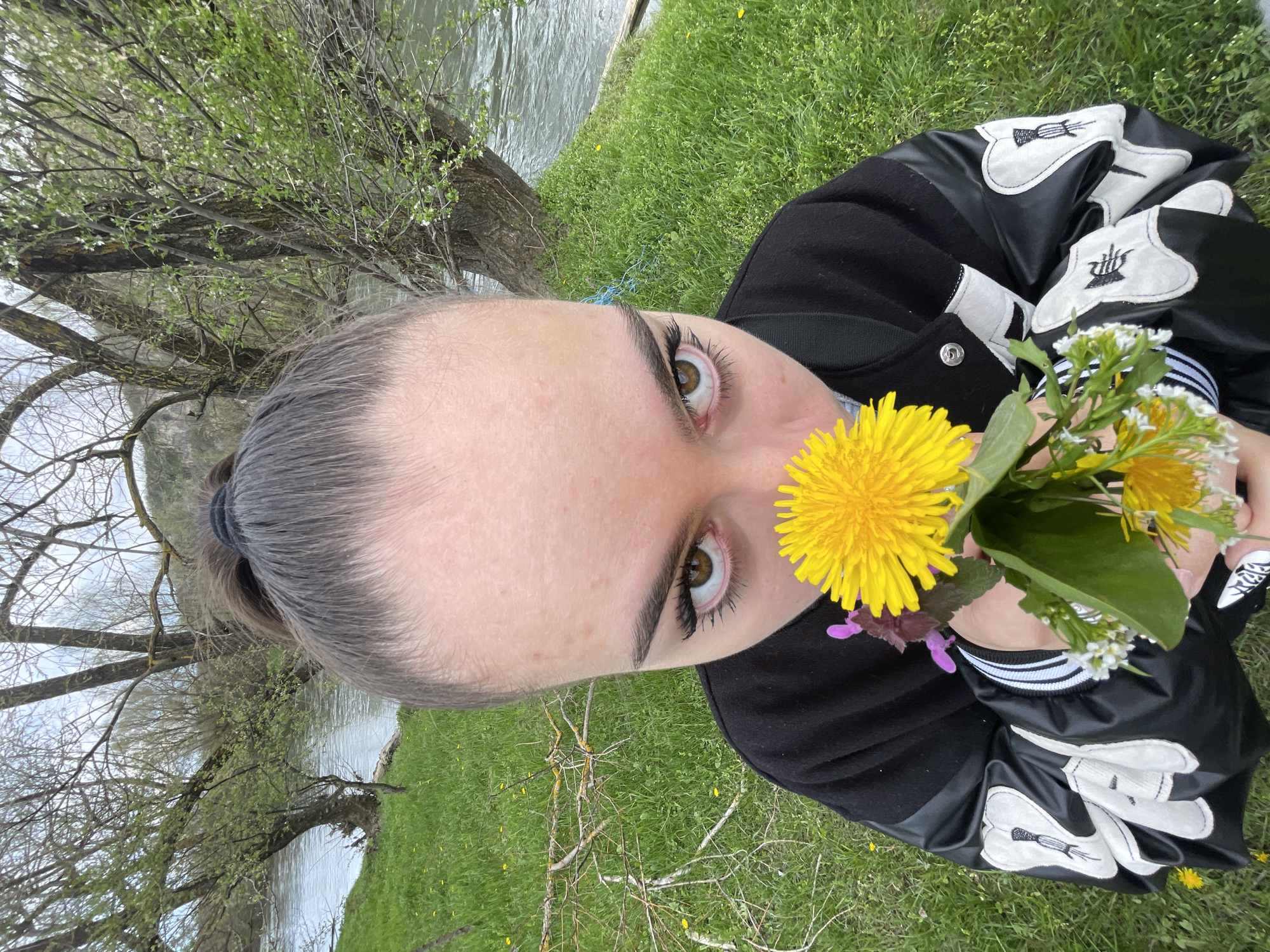 A young person with brown eyes and dark eyelashes is holding a small bouquet of dandelions and white wildflowers in front of their face. They are wearing a black bomber jacket with white designs on the sleeves. The background is a grassy area with trees and a body of water. The photograph appears to have been taken at a low angle, tilted slightly. The location is not discernible as Barkach, Bulgaria from the details in the image.