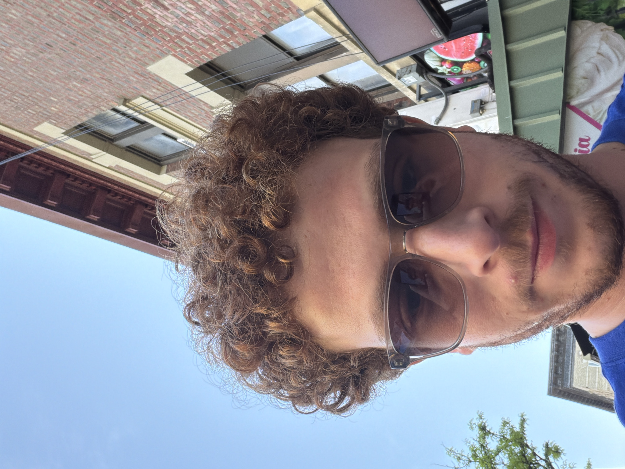 The image is a close-up selfie of a man with curly brown hair and a beard. He is wearing sunglasses. Behind him are buildings, including a brick building with windows and another building with green trim. The sky is visible in the background. It's not possible to determine the location with certainty based on the provided visual information.