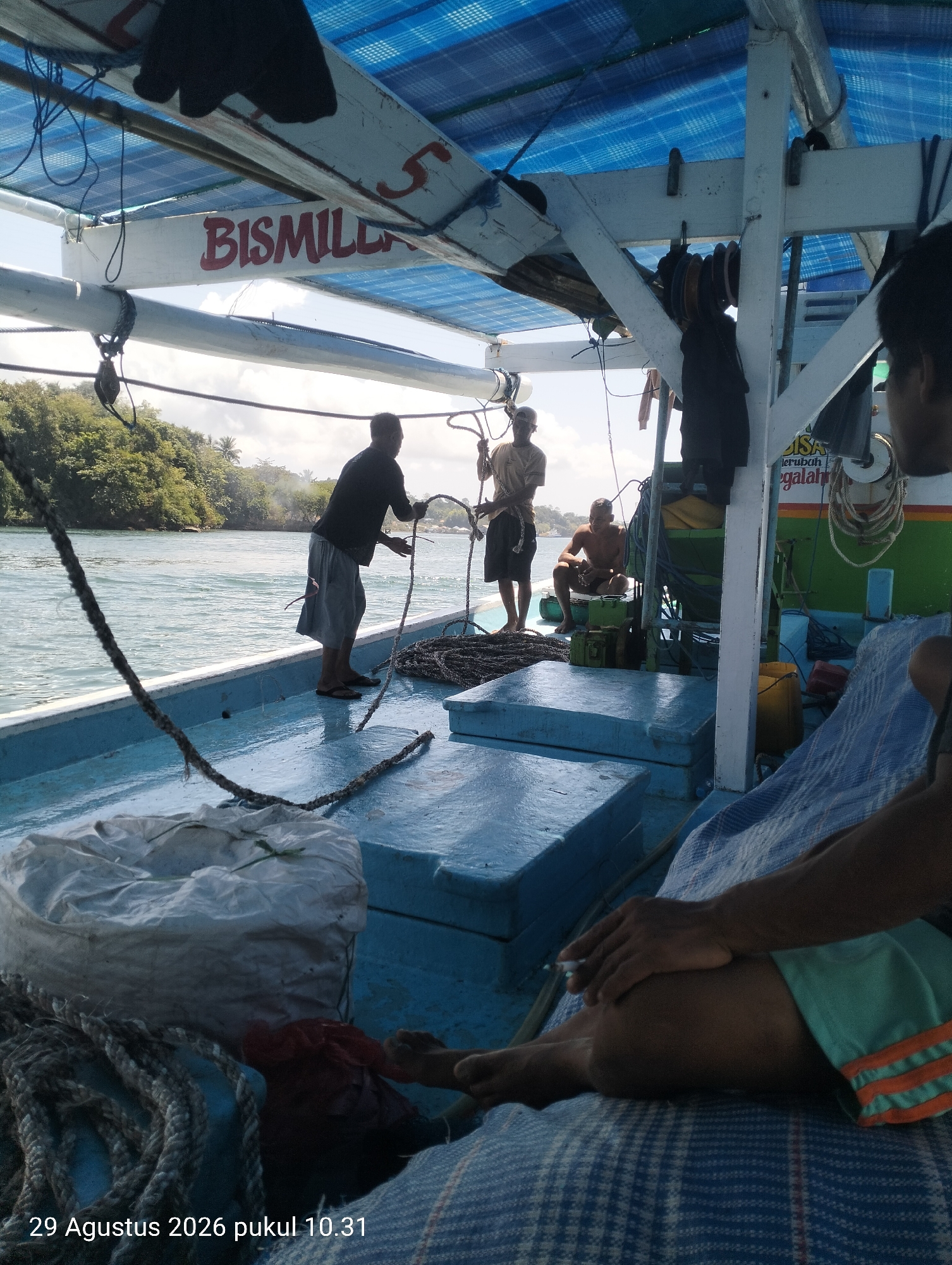 The image shows a view from the deck of a blue boat, likely a fishing vessel, on a body of water. The boat has a blue tarp overhead supported by white wooden beams. A white beam on the port side has the word "BISMILLAH" painted in red. Three men are visible on the boat. One man, wearing a dark long-sleeved shirt and grey shorts, is standing near the port railing and appears to be handling a rope. Another man, wearing a light brown t-shirt and dark shorts, is standing further aft, also near the railing. A third man is seated on the deck in the background. The water is calm and appears to be a river or bay, with lush green vegetation and trees visible on a distant shoreline. The overall scene suggests a working vessel in a tropical environment. The date and time stamp at the bottom left of the image indicates "29 Agustus 2026 pukul 10.31" which translates to "August 29, 2026 at 10:31 AM". The location is identified as Kendari, Indonesia.