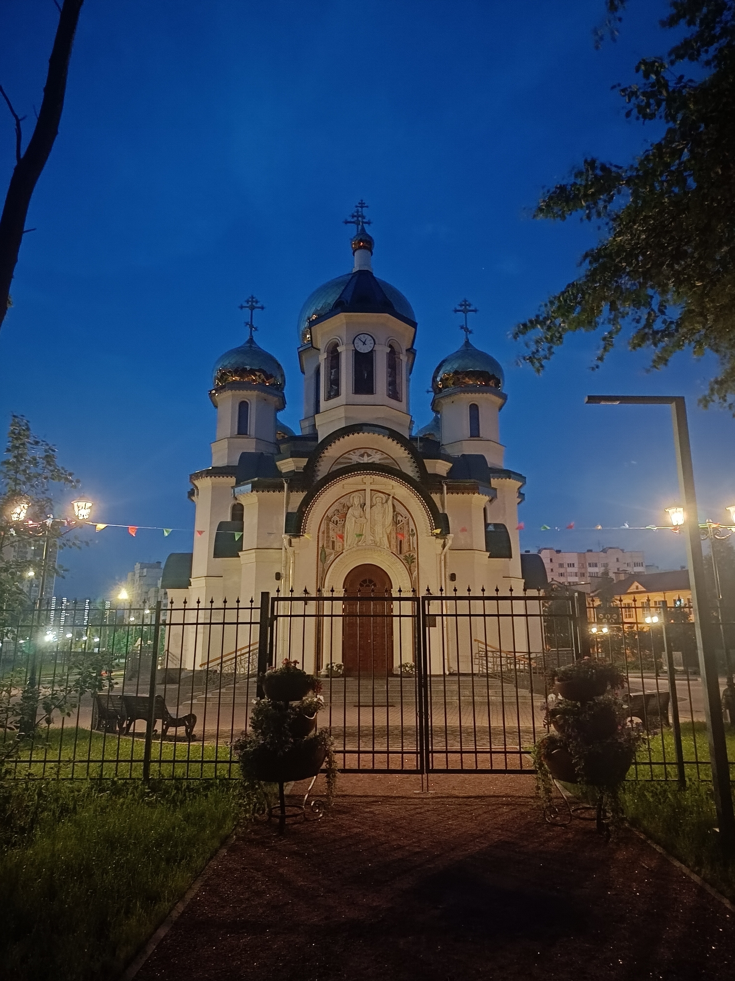 The image depicts the Church of the Icon of the Mother of God of All the Afflicted in Saint Petersburg, Russia, at dusk. The church, a prominent landmark, features multiple domes, intricate facade details, and is illuminated by artificial lights, highlighting its architectural grandeur against the deep blue twilight sky. A black metal fence encloses the church grounds, separating the viewer from the building. Inside the fenced area, a pathway leads towards the entrance, flanked by manicured grass and decorative planters. Benches are visible within the churchyard. In the background, apartment buildings and streetlights are discernible, indicating the urban setting. The scene is serene and peaceful, capturing the church as a beacon in the darkening city.