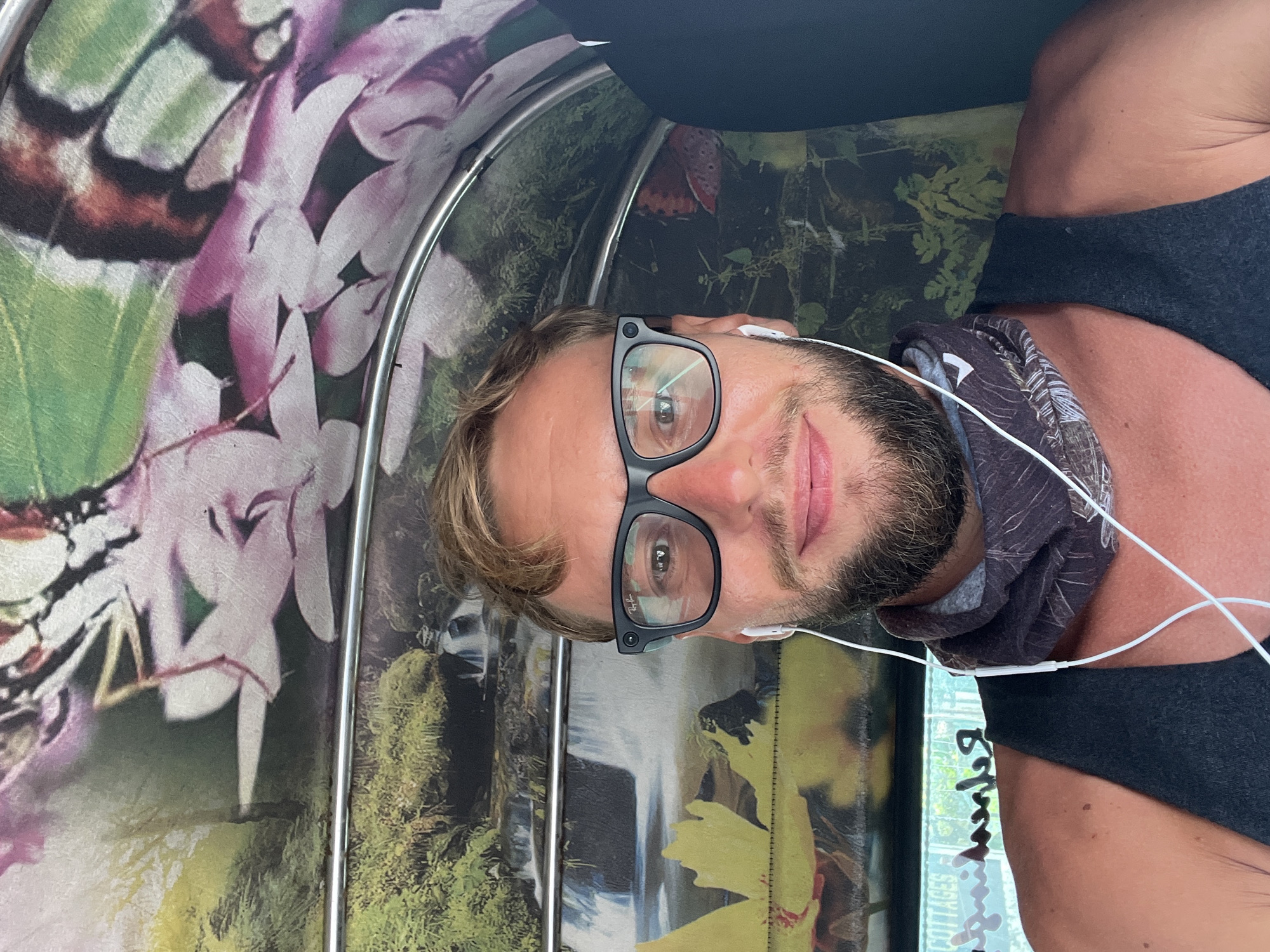 A man with blond hair, a beard, and glasses is taking a selfie in a tuk-tuk. The interior of the tuk-tuk is decorated with vibrant murals of nature scenes, including waterfalls, lush greenery, and butterflies. The man is wearing a black tank top and a patterned neck gaiter. White earbuds are visible. In the background, through the open side of the tuk-tuk, text that appears to read "COTTAGE" is visible.