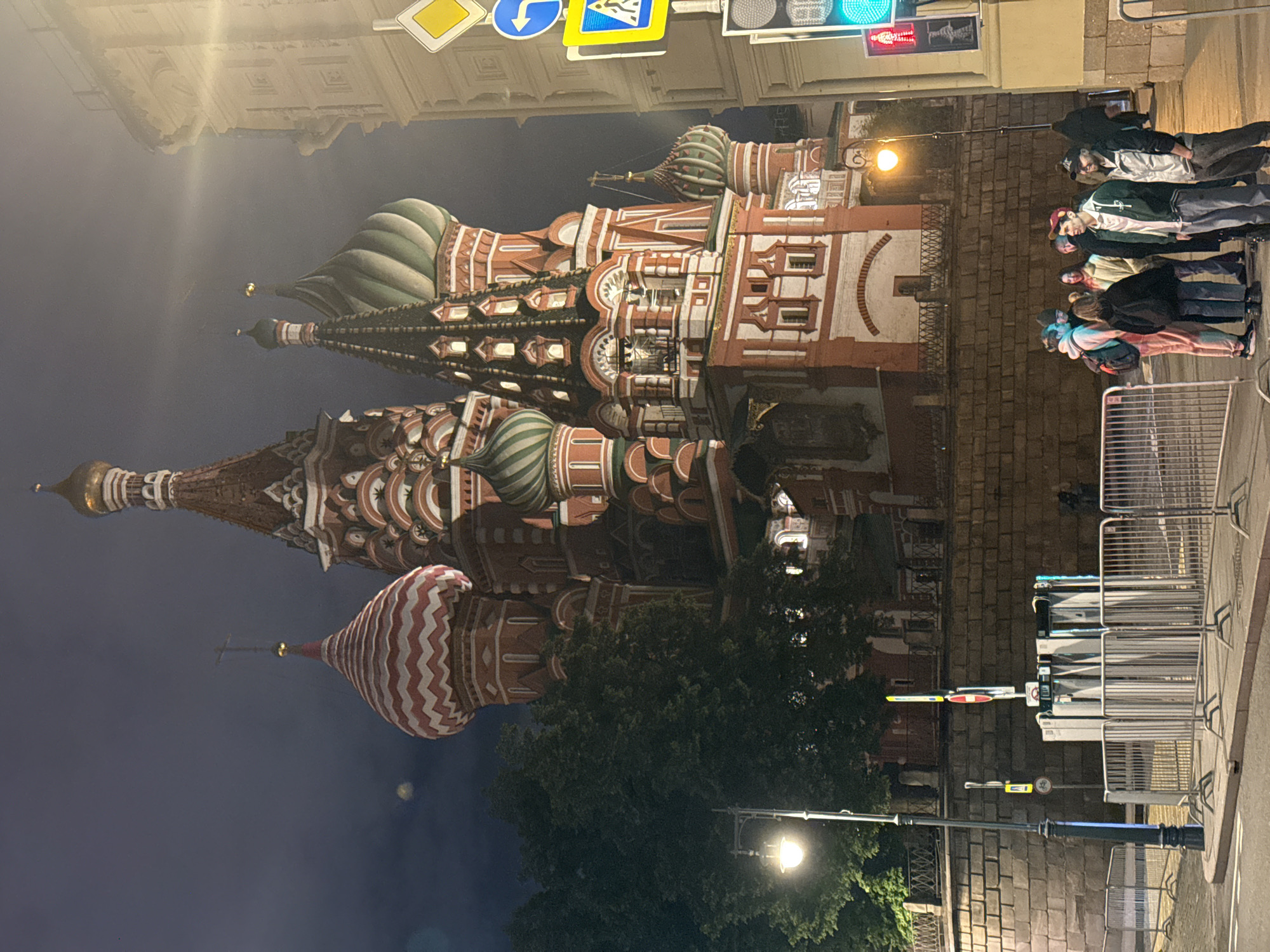 The image shows St. Basil's Cathedral in Moscow, Russia, at night. The cathedral, a multi-domed structure with distinctive onion domes and colorful, ornate details, is the main subject. In the foreground are several people standing on a sidewalk behind a row of metal barriers. A street lamp is visible next to the barriers. Street signs and traffic lights are above the cathedral. The sky is dark.