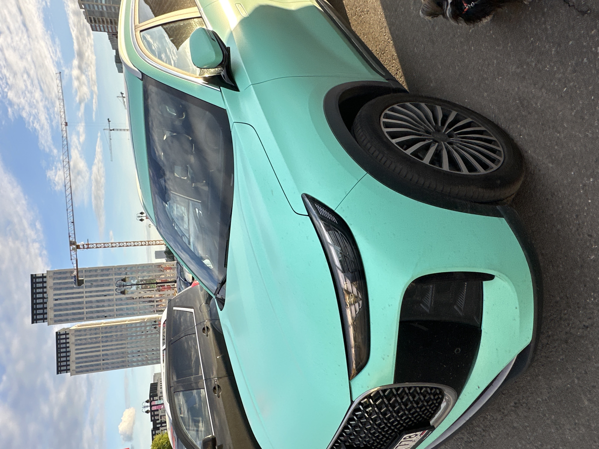 The image, rotated 90 degrees counter-clockwise, captures the front left portion of a distinctive mint green car, possibly a crossover or SUV, parked on an asphalt surface. The vehicle features a modern design with sleek, dark-tinted headlights and a textured front grille. Its multi-spoke wheel and tire are clearly visible on the pavement. In the upper right corner of the correctly oriented image, a small, dark-furred dog on a red leash is partially visible near the car's front wheel.
Another dark-colored vehicle, likely a van or a larger SUV, is parked closely to the left of the mint green car. The background reveals a bright blue sky dotted with scattered white clouds. Several tall, modern high-rise buildings, characterized by numerous windows, dominate the distant skyline, signifying an urban environment. Towering construction cranes are also visible, indicating ongoing development in the area, consistent with scenes in Moscow, Russia. Streetlights can be seen between the buildings.
The scene is brightly lit by natural sunlight, suggesting it is daytime, likely late morning or early afternoon, under clear weather conditions. A small, partially obscured license plate is visible on the front of the mint green car, showing what appear to be Cyrillic characters, which aligns with the location context of Moscow, Russia. The primary activity depicted is parking, with no human figures explicitly visible, though the dog suggests human presence nearby.