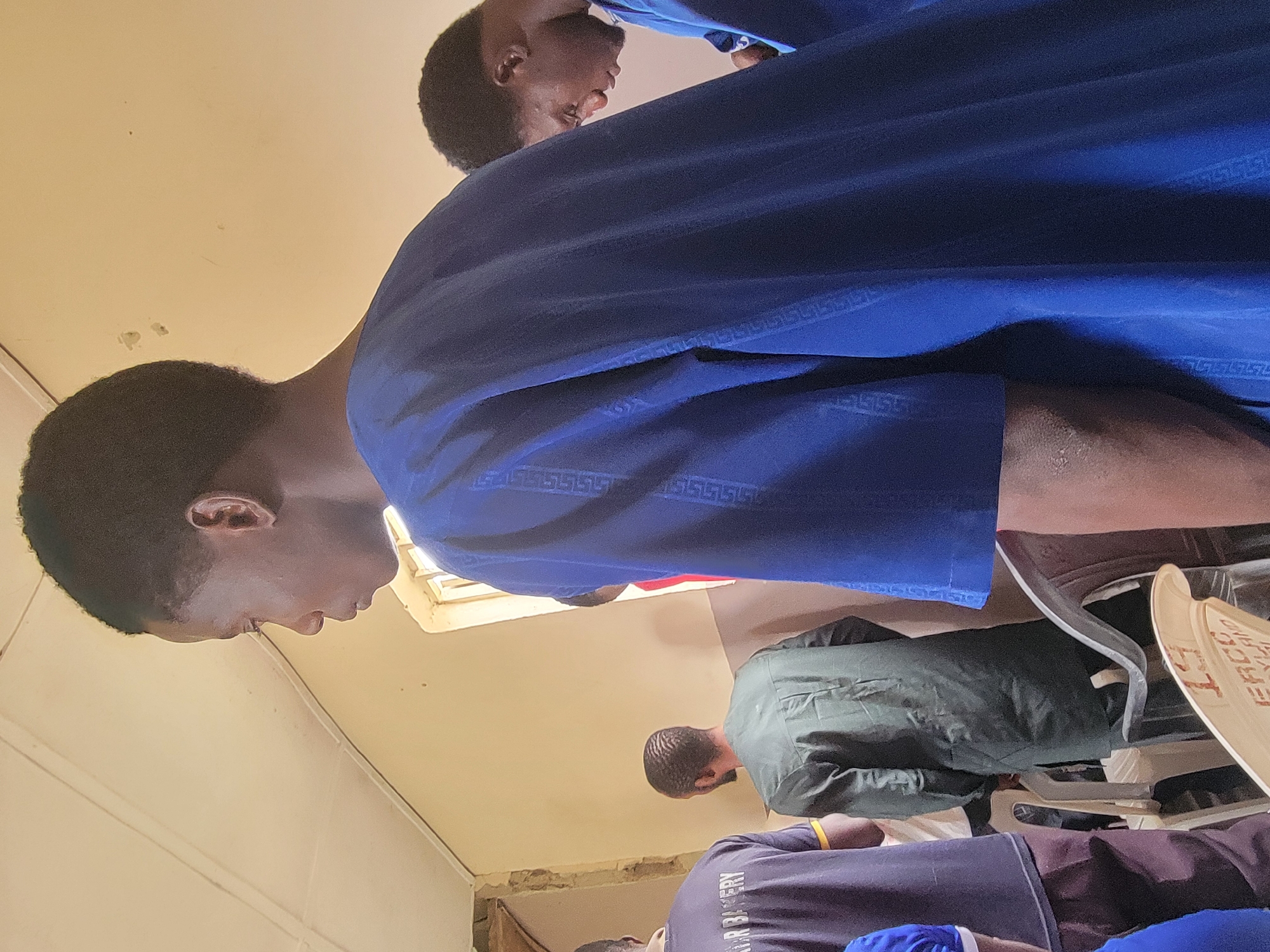 A high-angle shot of three people, with only the top portions of each figure visible. The closest individual is wearing a blue garment with a geometric border pattern. In the center, a person wears a dark green jacket. The third person, at the bottom of the frame, is wearing a purple shirt with the word "BAKERY" visible. A window provides light into the room. The location is Madalla, Nigeria, based on the text visible on a light-colored object on the right side of the image.