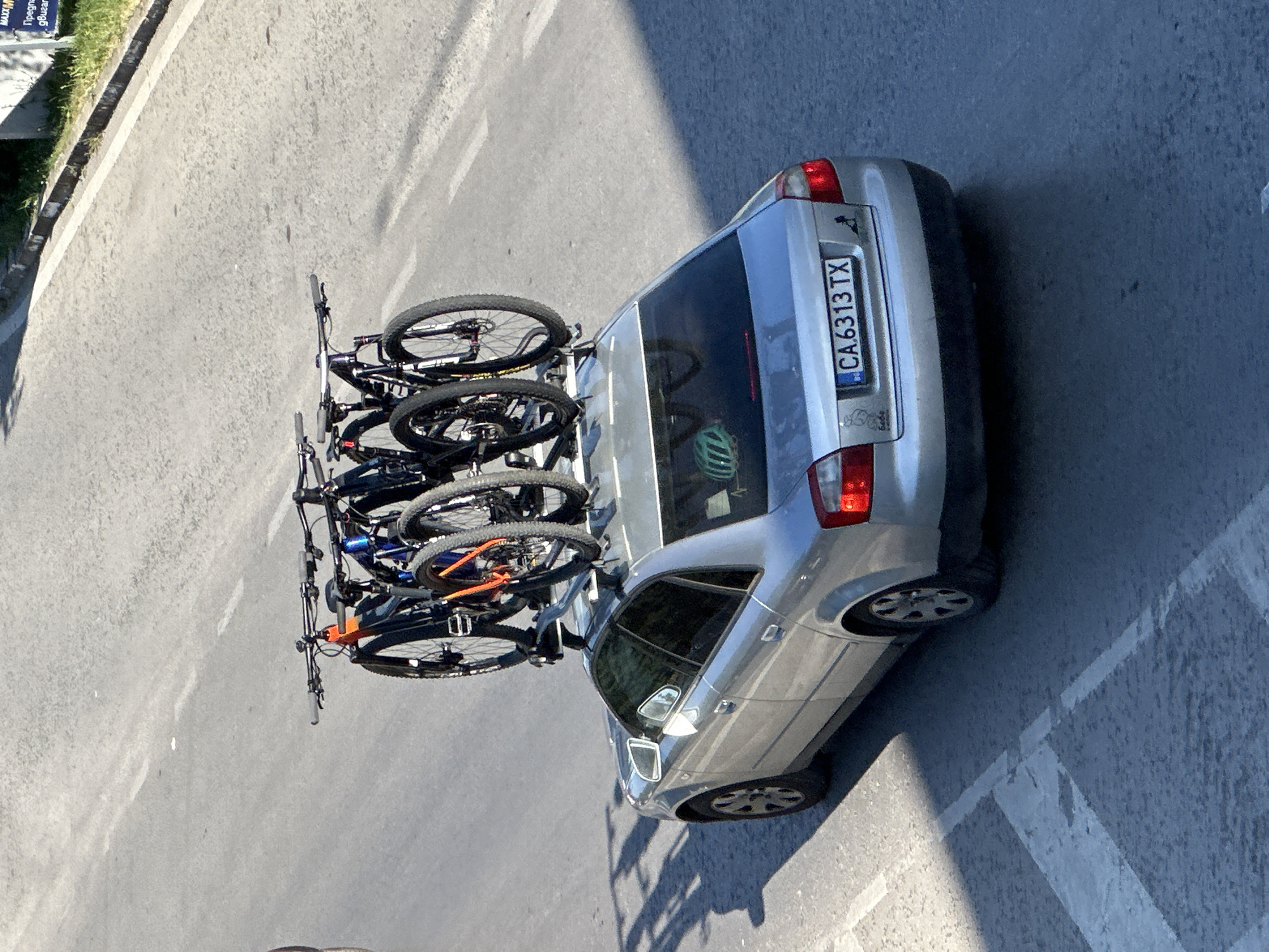 A silver sedan is parked on a street, possibly in Veliko Tarnovo, Bulgaria. The car is equipped with a roof rack carrying three bicycles. The license plate reads "CA 6313 TX". A mint green bicycle helmet is visible inside the car through the rear window. The street is paved and appears to be a sunny day, casting shadows on the asphalt. A white line marking is present on the road. A blurred sign with text is visible in the background to the right.