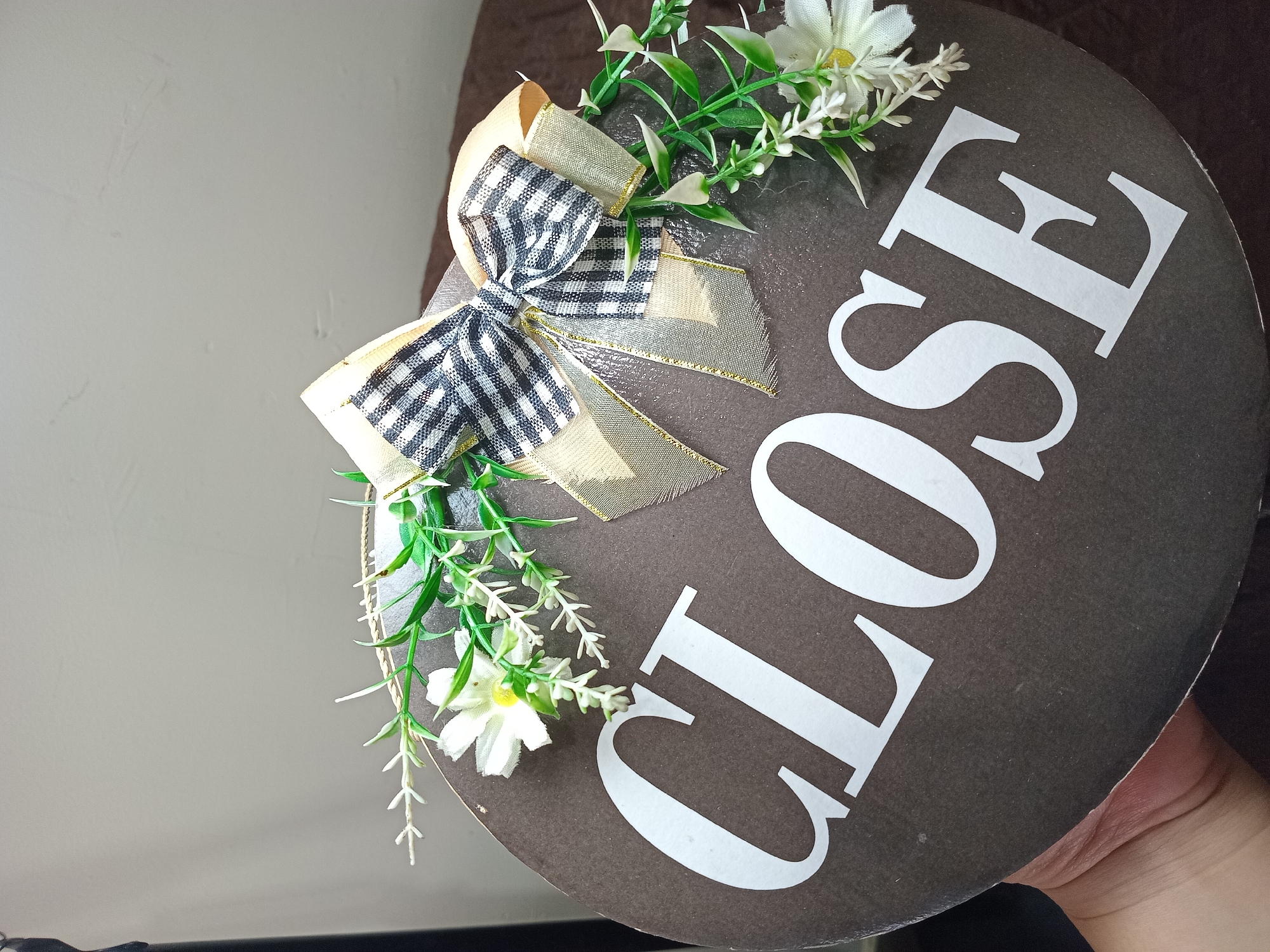 A round decorative sign, predominantly dark gray or brown, is displayed, featuring the word "CLOSE" in large, white, sans-serif capital letters on its right side. The sign is adorned with a decorative bow and artificial floral elements in its upper left quadrant. The bow is multi-layered, consisting of a black and white gingham patterned ribbon tied over a wider, sheer gold ribbon with golden edges. Clusters of artificial white flowers and green leafy foliage are arranged around and beneath the bow.
The sign is held by a hand visible in the bottom right corner, suggesting it is being presented or moved. The background behind the sign includes a light-colored, possibly off-white, wall on the left and a darker, soft fabric or surface on the right. The overall impression is that of a handcrafted or customized business sign, indicating that a shop or establishment is currently not open. The image provides no specific time of day or weather cues, and the location context is North Kuta, Indonesia.