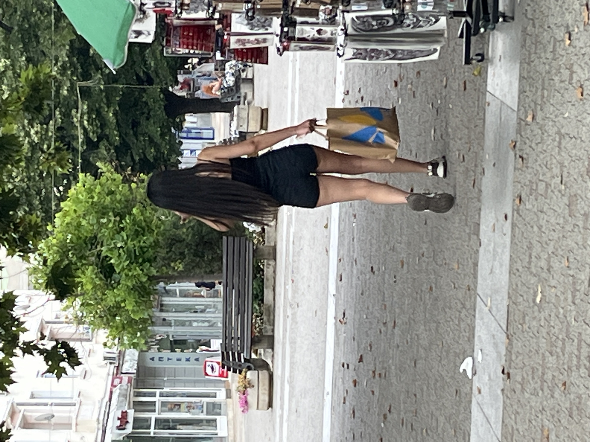 The image shows a person with long dark hair wearing a black t-shirt and black shorts, walking on a sidewalk in Sliven, Bulgaria. They are carrying a brown paper bag with a blue and yellow design. The person is walking away from the camera, and their legs and feet are visible. They are wearing dark sneakers.
The scene appears to be a street with buildings on the left side and trees and shops on the right. There is a black bench on the left side of the sidewalk, and some flowers in a planter near the bench. The ground is paved with concrete and scattered with fallen leaves or debris. The buildings have multiple windows and some signage. In the background, there are displays of various items, possibly from a market or shops. A green flag is visible in the upper left corner.
The lighting suggests it is daytime, likely a bright day given the shadows. No specific emotions are evident due to the person's pose and distance. The overall impression is of a casual walk on a street, possibly after shopping.
Visible text includes "AUTEXA" on a sign above a shop window. There are also other signs with text that is too small or blurry to read.