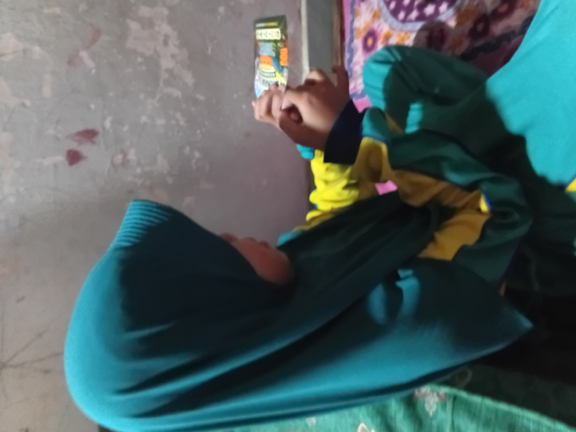 A young person wearing a teal hijab and a yellow and teal jacket is sitting and looking at a phone. The background shows a rough, textured wall with some reddish marks and a patterned blanket or rug on the floor. The scene appears to be indoors in a residential setting, possibly in Plemahan, Indonesia.