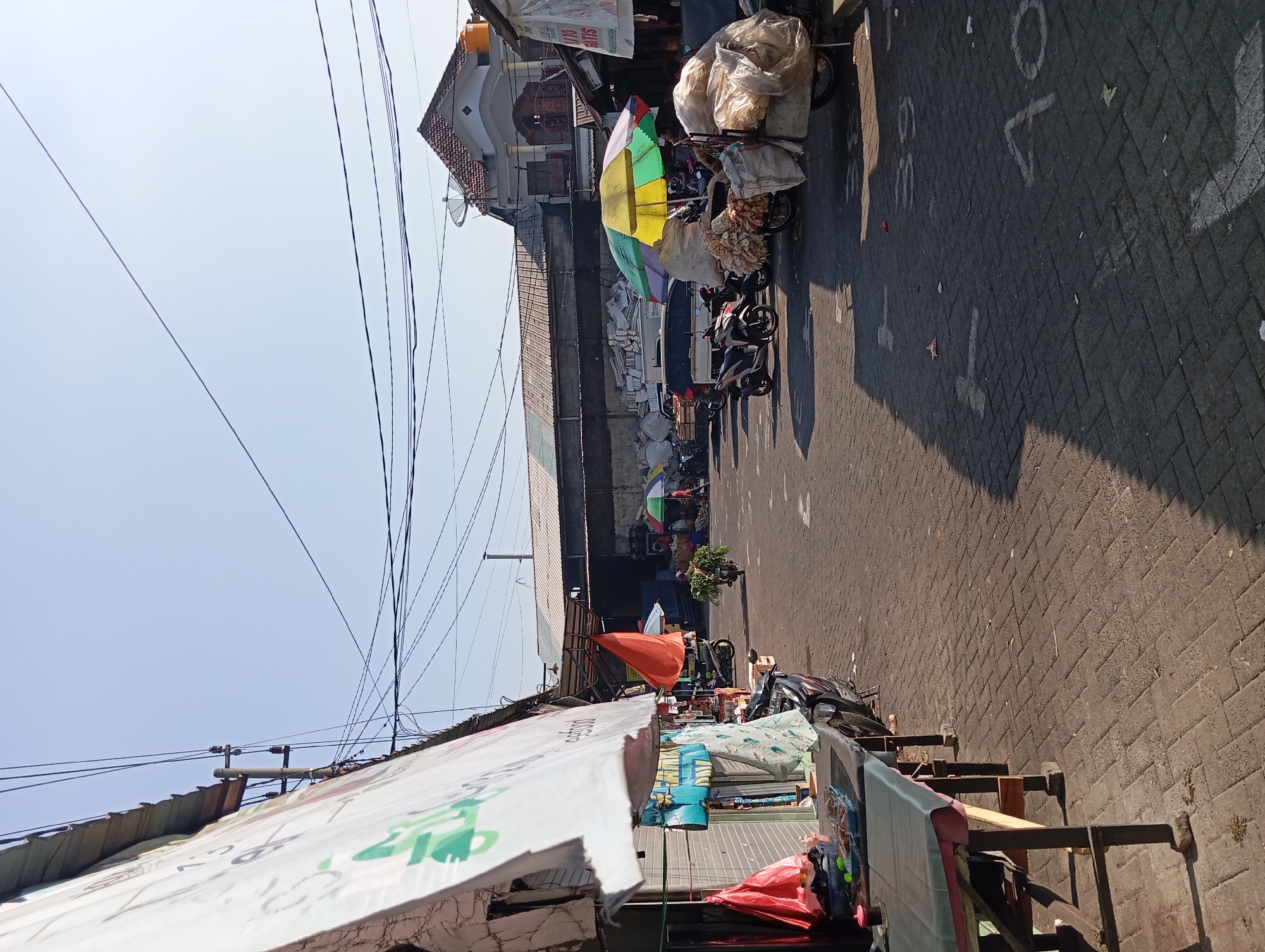 The image depicts a street market in Kepulungan, Indonesia, on a clear, sunny day. The foreground is dominated by a market stall on the left, featuring a table covered with a dark green cloth. Several items are visible on the table, including plastic bags, baskets, and containers, suggesting it is set up for selling goods. A motorcycle is parked next to the stall.
The street itself is paved with interlocking bricks. Several other market stalls and vendors are scattered throughout the scene. To the right, more motorcycles are parked, some with goods loaded onto them. Several umbrellas, including a prominent yellow and green one, provide shade for vendors and their products. There are also bags of goods, possibly produce or other commodities, stacked on or near the motorcycles.
In the background, a long, low building with a corrugated metal roof stretches across the frame. Beyond that, a residential building with a tiled roof and a satellite dish is visible. Power lines crisscross the sky, typical of urban or semi-urban environments. A few people are present in the market, some tending to their stalls or goods, and one individual is seen riding a motorcycle laden with leafy green vegetables. The overall atmosphere suggests a typical bustling market scene under the bright sun.