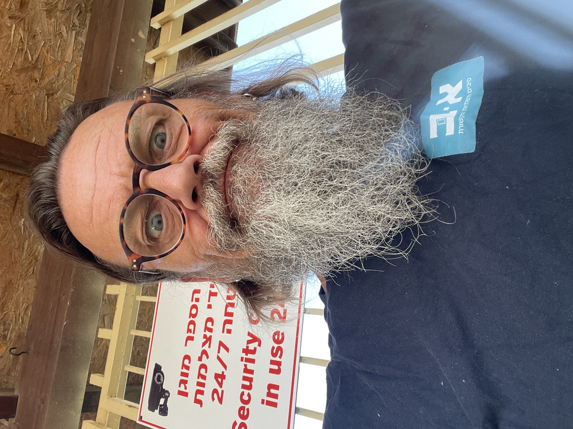 In a selfie taken outdoors, a man with a long gray beard and round glasses looks directly at the camera with wide eyes. He is wearing a dark blue t-shirt with a light blue logo on the left side of his chest. Behind him, a white sign with red lettering and a black icon of a security camera is visible. The sign appears to be a "Security in use 24/7" sign. The background consists of wooden structures and what appears to be a fence. The setting is the city of Even Yehuda, Israel.