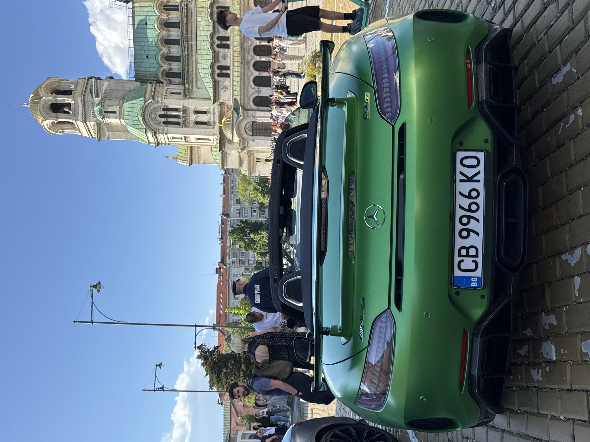 The image shows a green Mercedes-AMG GTR Roadster parked on a brick-paved area. The car's license plate reads "CB 9966 KO," indicating it is registered in Bulgaria. The back of the car is facing the camera, and the top is down. The Alexander Nevsky Cathedral in Sofia, Bulgaria, is visible in the background, confirming the location. Several people are nearby, including some standing on the far side of the street and two people standing behind the car. A light post is visible on the left side of the frame. The sky is blue with some clouds.