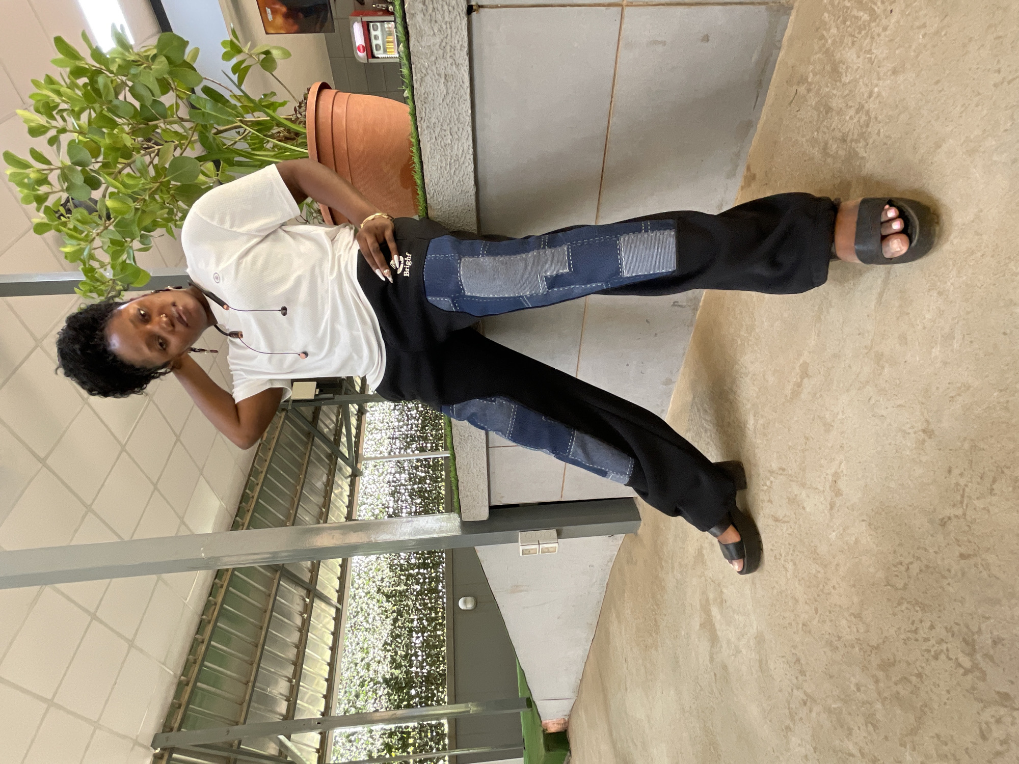 A young woman is captured in a full-body shot, posing in what appears to be a covered outdoor or semi-indoor area in Abuja, Nigeria. She has dark skin and dark, curly hair, and looks directly at the camera with a neutral expression. She is wearing a white short-sleeved t-shirt with a subtle dark logo on the chest, paired with black wide-leg trousers. The trousers feature distinct blue and lighter blue denim-like patches of varying shapes and patterns on the front of both legs, some with visible white stitching. On her right hip, the word "Bright" is visible in white script on her black pants. She accessorizes with a gold bracelet on her right wrist, and white earbuds with orange accents are visible. She wears black open-toed slides on her feet, revealing white-painted toenails.
She stands with her weight slightly shifted, leaning back against a light gray concrete pillar or wall. Her left hand is raised, resting near her head, while her right hand is on her hip. To her right, a large potted plant with abundant green foliage sits in a terracotta-colored pot. The background features a textured light beige floor and a ceiling with white square panels. Beyond the pillar and plant, there's a structure with a corrugated metal roof and a lush, green artificial hedge or wall, suggesting an open-air but sheltered environment. Metal support beams are also visible in the background. The lighting indicates it is daytime.