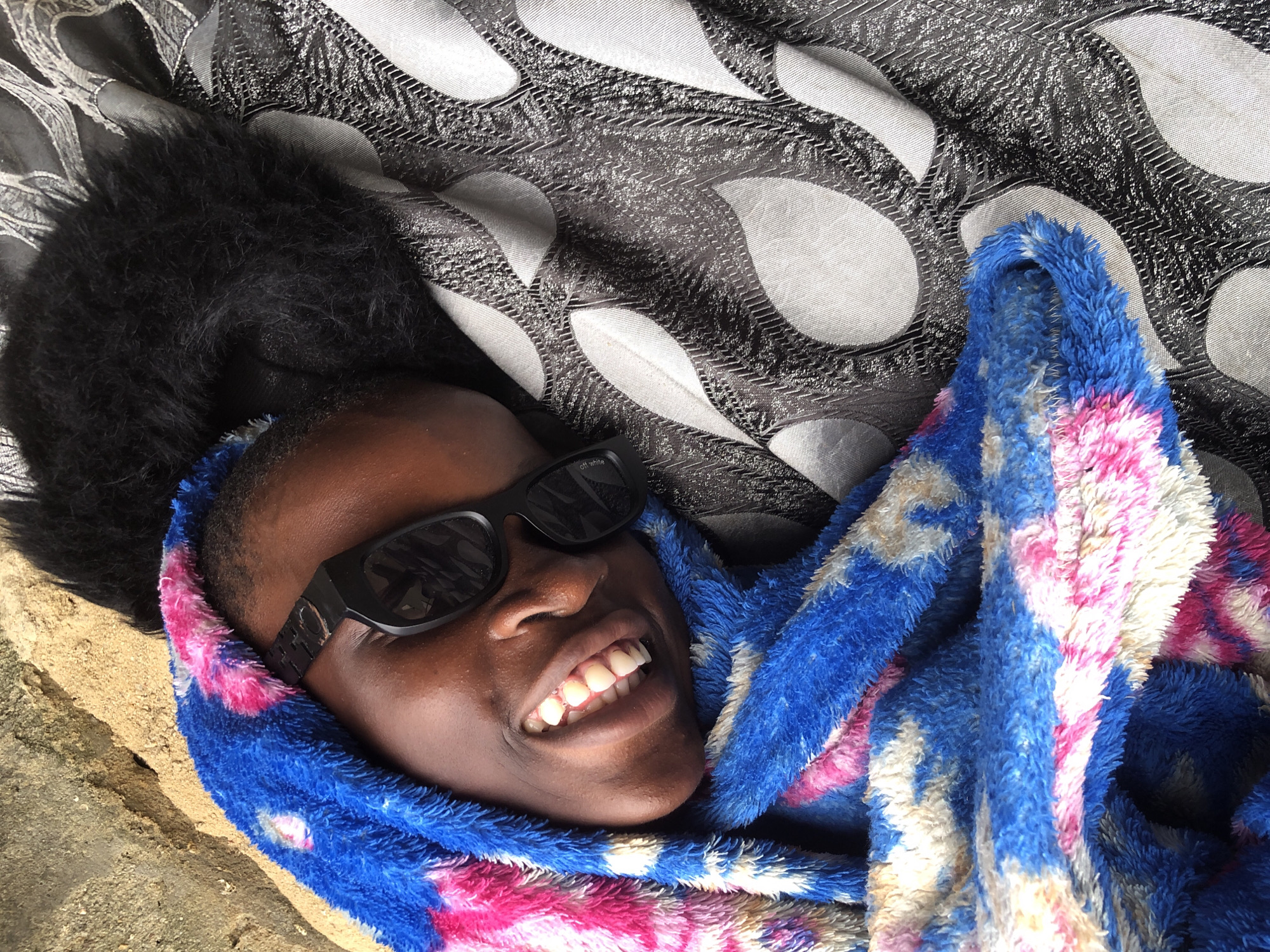 In Ado, Nigeria, a close-up shot shows a Black child lying down and wrapped in a blue, pink, and white patterned fuzzy blanket. The child wears black sunglasses and has their mouth open, revealing teeth, and appears to be smiling. Their dark, fuzzy hair is visible at the top of their head. The child is positioned on a gray textured fabric with an oval pattern. To the left of the child, a sandy, light-colored surface is visible, possibly a wall or ground.