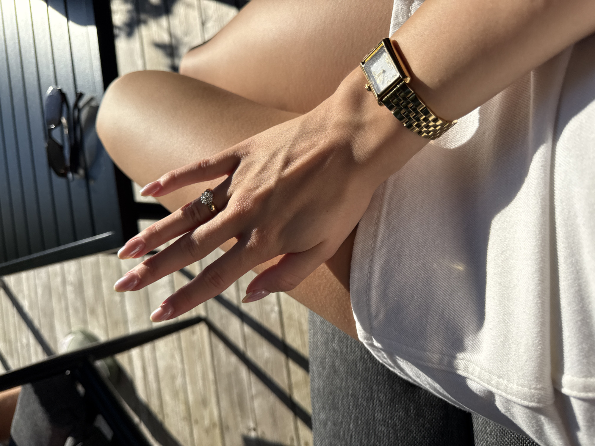 The image shows a close-up of a person's hands and legs. The person is wearing a gold watch on their left wrist and a ring on their right ring finger. The background is blurred, but appears to be an outdoor setting with wooden decking and furniture. The lighting suggests it is daytime. The location is identified as Harstad, Norway.