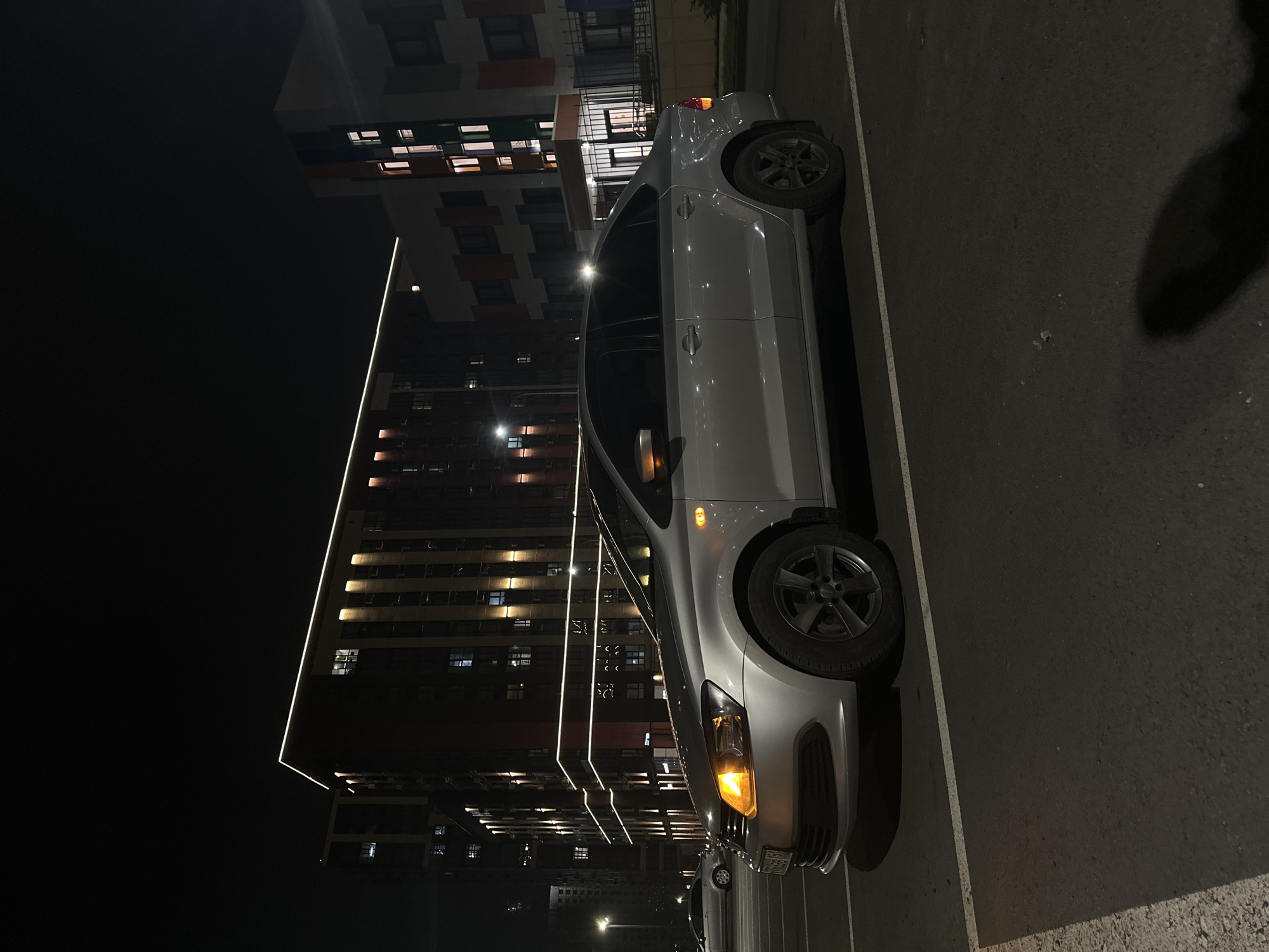 The image displays a nighttime urban scene in Rostov-on-Don, Russia. In the foreground, a silver four-door Volkswagen Polo sedan is parked, angled slightly left, within a marked asphalt parking space. The vehicle's front left headlight and turn signal are active, emitting an orange-yellow light, as is a side marker light on the left fender. A Russian license plate is affixed to the front bumper. The car is equipped with dark, multi-spoke alloy wheels.
In the mid-ground, a tall, multi-story building with a dark facade dominates the left and center. It is distinctively illuminated by horizontal white LED strip lighting along its roofline and possibly lower architectural bands, complemented by vertical amber-colored lights on its facade. Numerous windows in this building are lit. To the right, a second multi-story building with a lighter-colored facade features a pattern of white, orange, blue, and green panels, also showing illuminated windows and a visible entrance with a dark fence. A second, lighter-colored sedan is partially visible, parked further behind the primary vehicle. Streetlights provide general ambient illumination. The sky is dark and clear. No individuals are present within the frame.