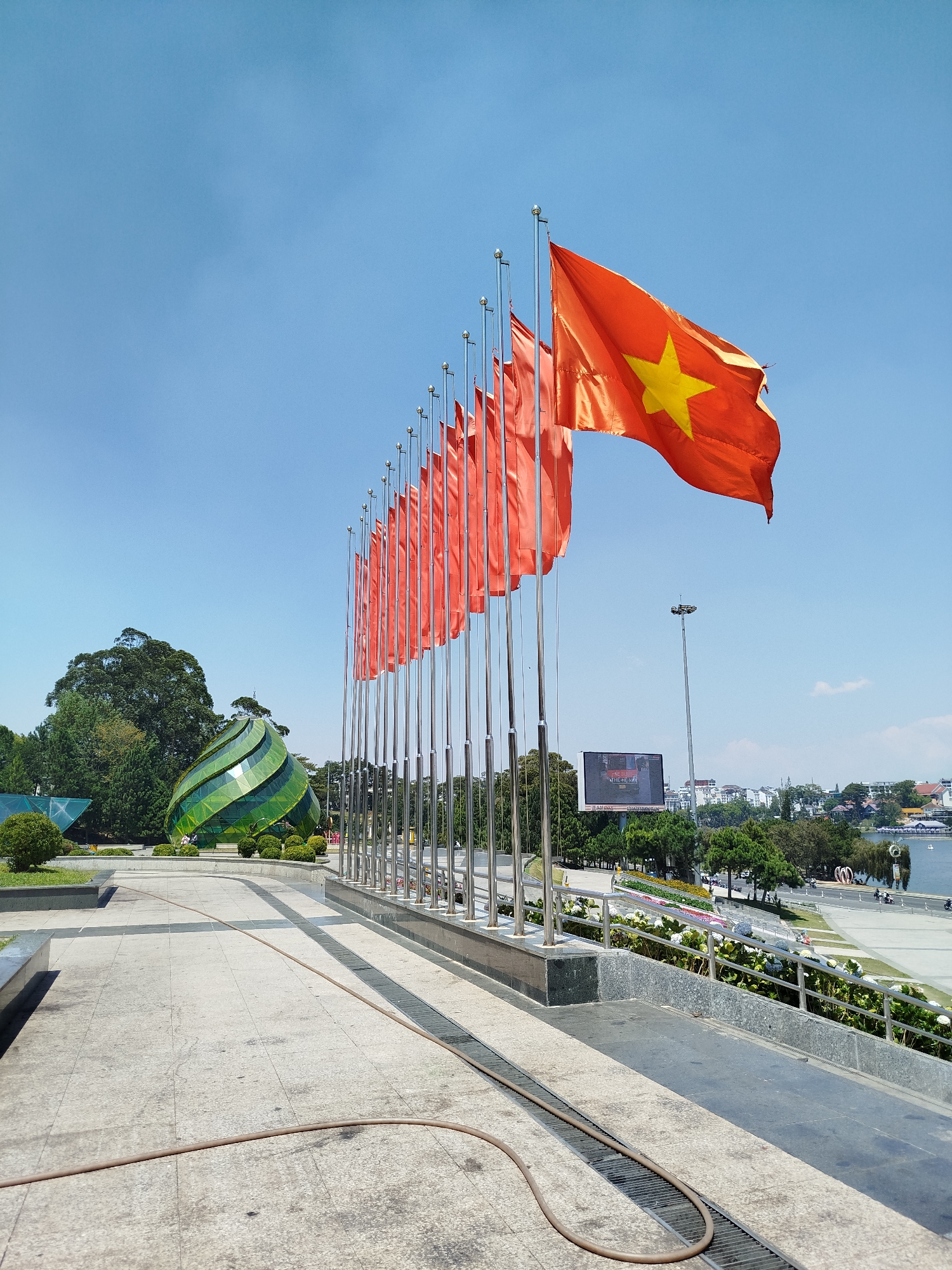 The image is a wide-angle shot taken in Da Lat, Vietnam, featuring a row of Vietnamese flags against a clear blue sky. The flags are orange with a yellow star and appear to be waving in the wind. The flagpoles are made of a shiny metal, likely stainless steel. In the background, to the left of the flags, is a modern green glass architectural structure resembling a water droplet or tear. Behind the flagpoles, there is an open space with trees, roads, and buildings. In the far background is a body of water. The foreground includes a paved walkway with a brown hose lying on the ground. The area appears to be a public square or plaza.