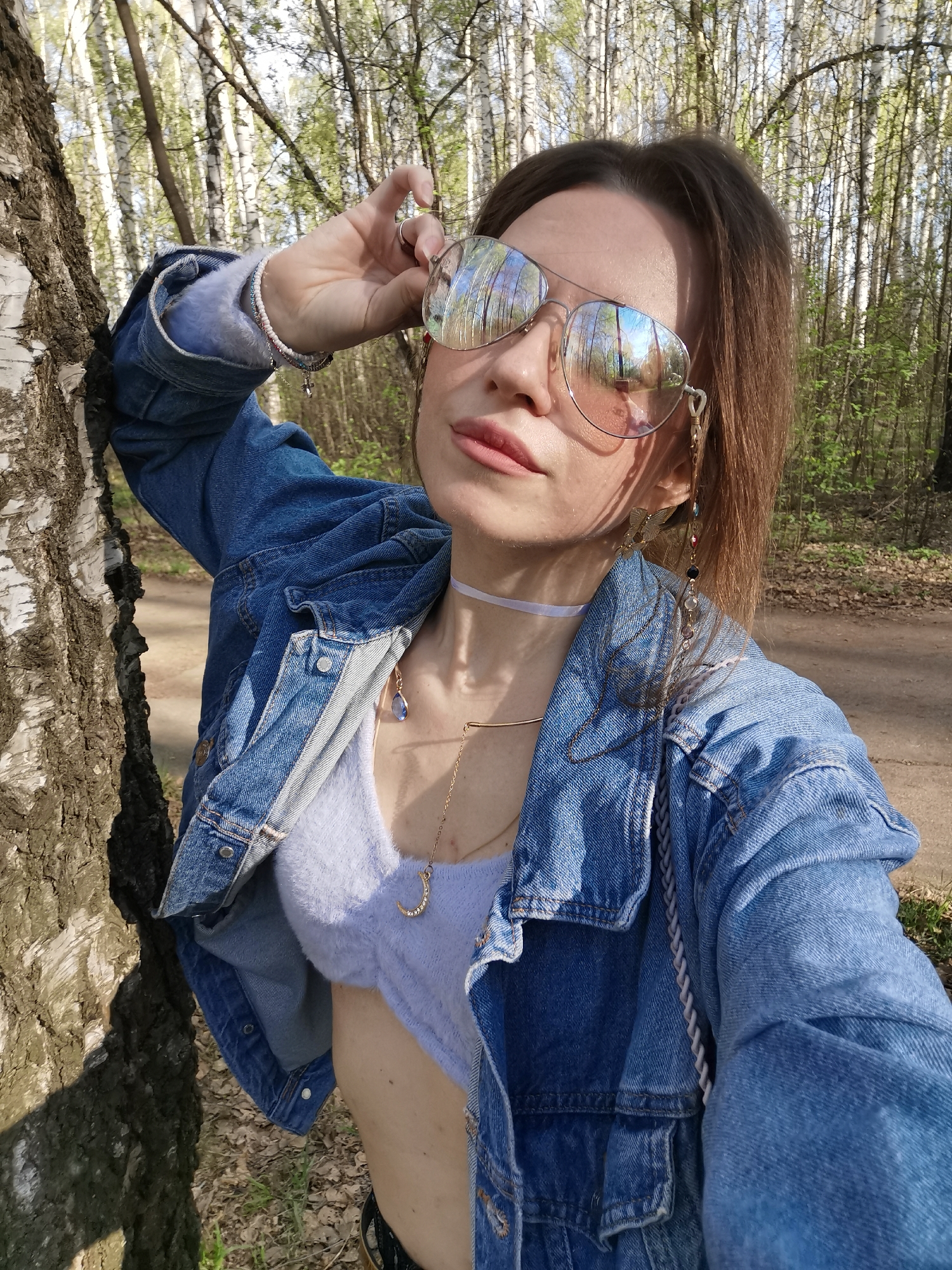 A woman stands in a forest, leaning against a birch tree. She is wearing mirrored aviator sunglasses, a denim jacket, a light purple top, and jewelry including a choker, necklace, and bracelet. Her hair is brown and partially pulled back. A dirt path is visible in the background, and there are many birch trees in the scene. The city location is unknown.