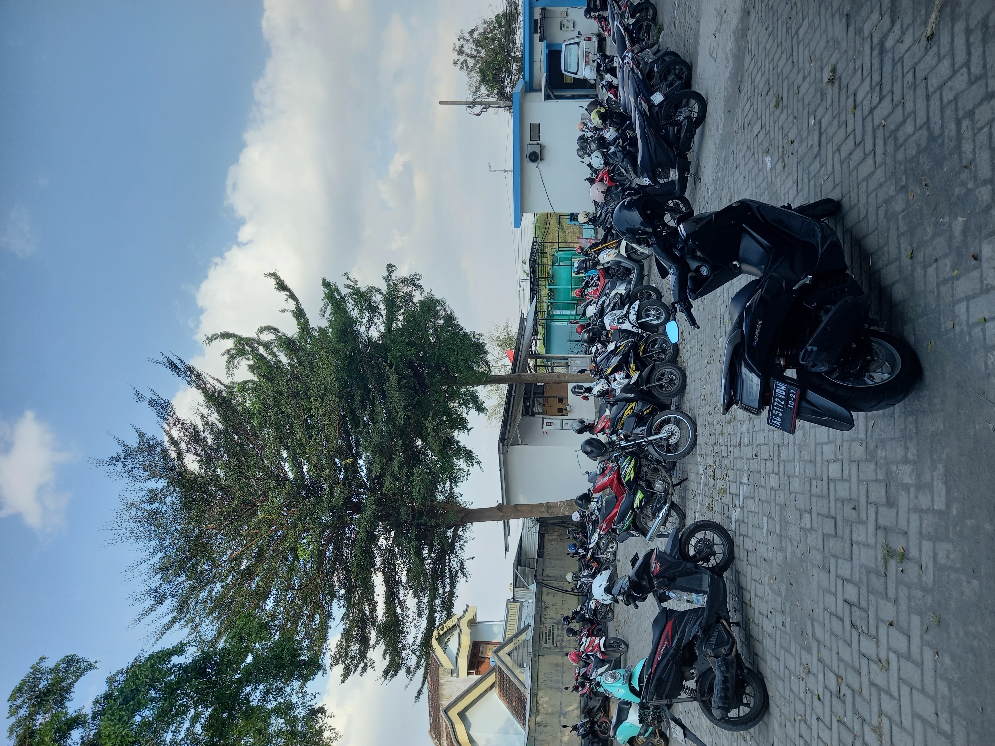 The image depicts a parking lot filled with numerous motorcycles, predominantly scooters, under a partly cloudy sky. The motorcycles are parked on a paved surface made of interlocking brick pavers. In the background, there are low-rise buildings, some appearing to be part of an industrial or institutional complex with white walls and blue trim. A prominent tree with dense green foliage stands in the mid-ground, casting shadows. A white van is visible parked near one of the buildings. The scene is likely a public or private parking area in Ploso, Indonesia, given the Indonesian license plate visible on one of the motorcycles. There are no people clearly visible in the image. The overall impression is of an outdoor, daytime scene in a built environment with a focus on the parked vehicles.