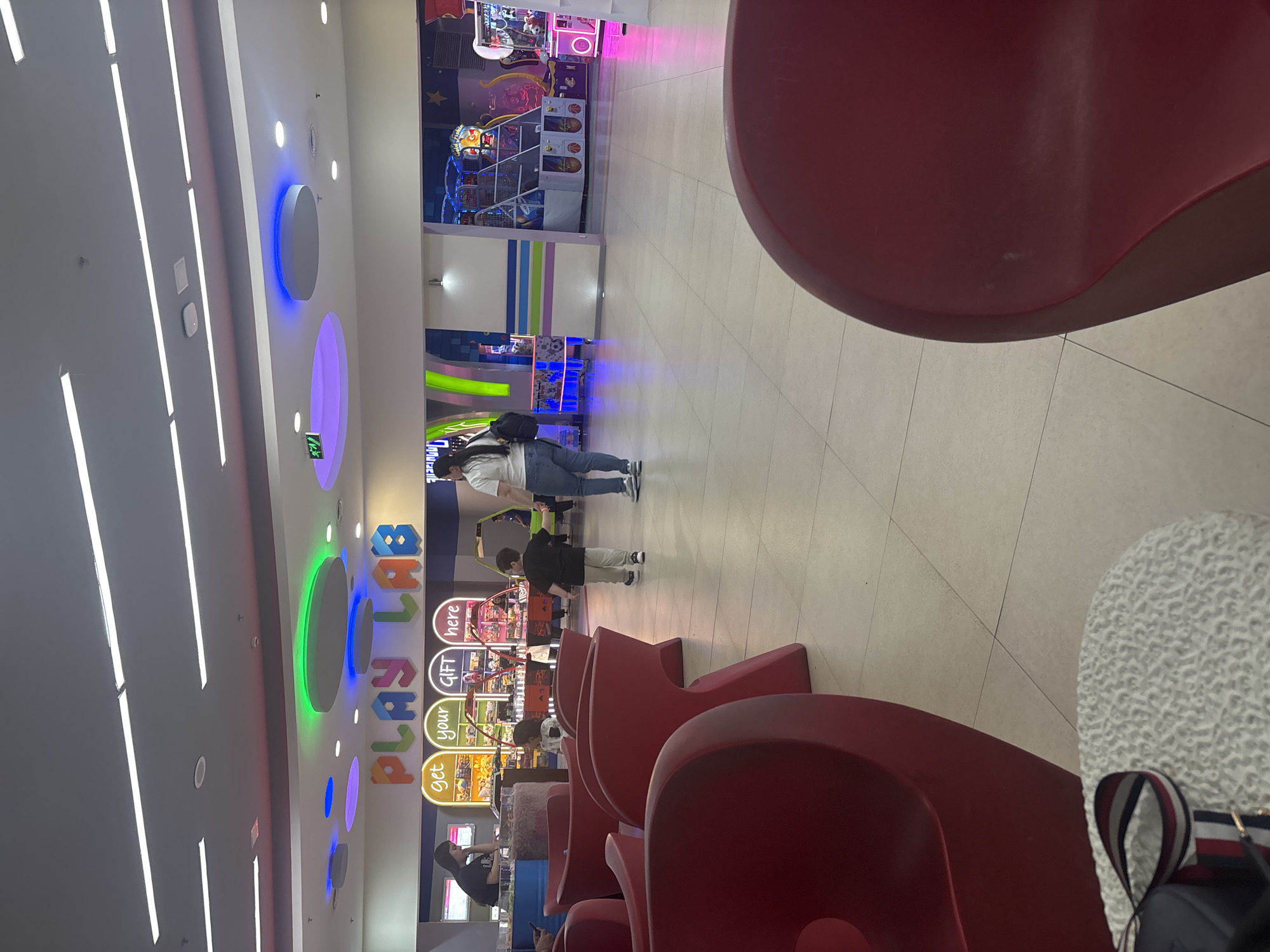 An indoor entertainment or play area, likely within a shopping mall in Yerevan, Armenia, is depicted in a portrait-oriented photograph. The space is brightly lit with artificial lighting, featuring long fluorescent strip lights on the ceiling alongside several circular lights that emit blue, purple, and green hues. The floor is tiled with light-colored square tiles.
In the foreground, a large, modern red chair is partially visible on the right, from which the photo appears to have been taken. Next to it on the floor is a white, textured object and a dark bag with a red, white, and blue striped strap. More similar red chairs are arranged in rows further back on the right, with a person sitting on one of them in the midground, looking towards the left.
On the left side of the frame, a prominent sign spells out "PLAY LAB" in colorful, block letters. Below it, a vertical stack of smaller, illuminated signs reads "get," "your," "GIFT," and "here," suggesting a redemption counter or prize area. An illuminated green "EXIT" sign with an arrow is also visible on the left wall.
In the midground, a woman wearing a white top and blue jeans, carrying a black backpack, stands facing away from the camera, looking towards the arcade games. A young child, possibly with her, is standing nearby in a dark top and light pants, seemingly interacting with or observing one of the arcade machines.
The background is filled with various arcade games, including basketball shooting games and claw machines, all brightly lit with colorful lights. The overall atmosphere suggests a lively and engaging environment for children and families. No specific time of day is discernible due to the indoor artificial lighting.
