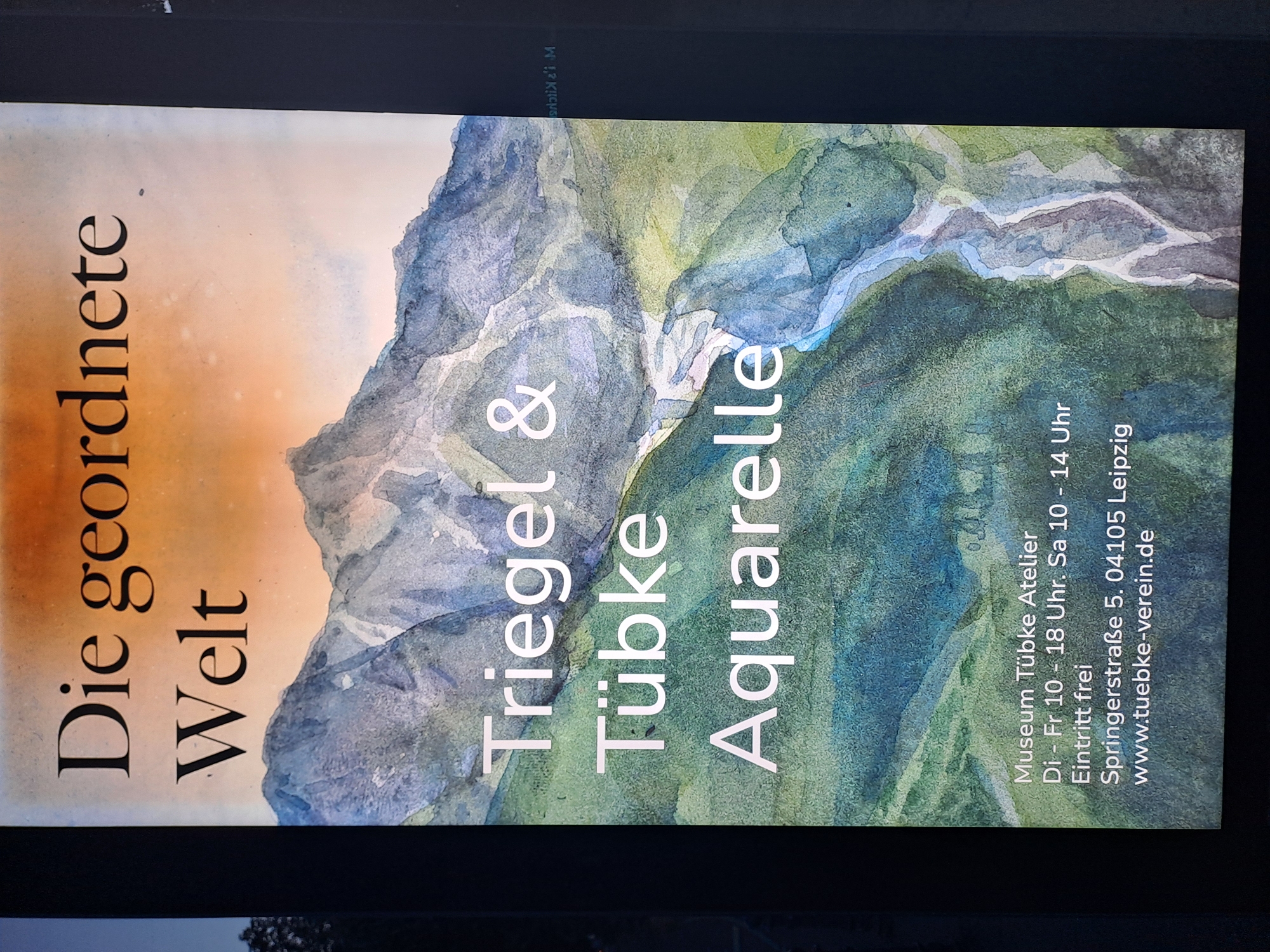 The image is a poster for an exhibition titled "Die geordnete Welt" (The Ordered World) by artists Triegel & Tübke, featuring watercolors. The exhibition is held at Museum Tübke Atelier, located at Springerstraße 5, 04105 Leipzig, Germany. The museum is open from Tuesday to Friday, 10:00 AM to 6:00 PM, and Saturday, 10:00 AM to 2:00 PM. Admission is free. The poster displays a watercolor painting of a mountainous landscape with a river flowing through it, with the text overlaid.