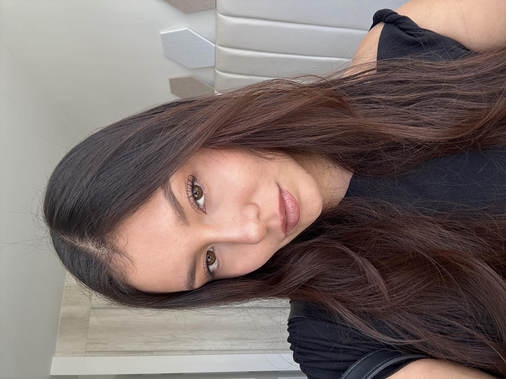 A young woman with long, dark brown hair is facing the camera with a slight tilt of her head. She has fair skin, brown eyes, and her lips are a neutral pink shade. She is wearing a black shirt. The background consists of a white wall with geometric patterns and a wooden surface. The location is not possible to be determined in this image and is likely not San Francisco Ocotlán, Mexico.