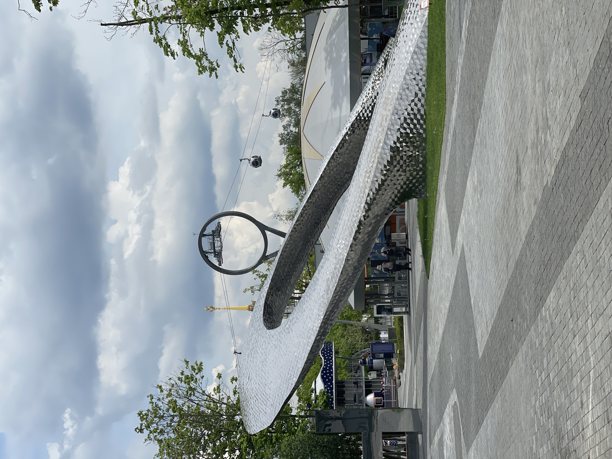 The image depicts a modern architectural landscape in an urban setting. In the foreground, there is a large, curving, metallic sculpture with a textured surface. A paved walkway stretches away from the sculpture, marked by patterns of grey stones. Adjacent to the walkway is a narrow strip of green grass.
In the background, there is a tall, circular structure with suspended gondola cars, likely part of a cable car system. Behind this, a building with a curved, white facade can be seen. People are visible near the base of the sculpture.
The sky is cloudy. The location appears to be a public park or recreational area, given the combination of art, infrastructure, and green space.
