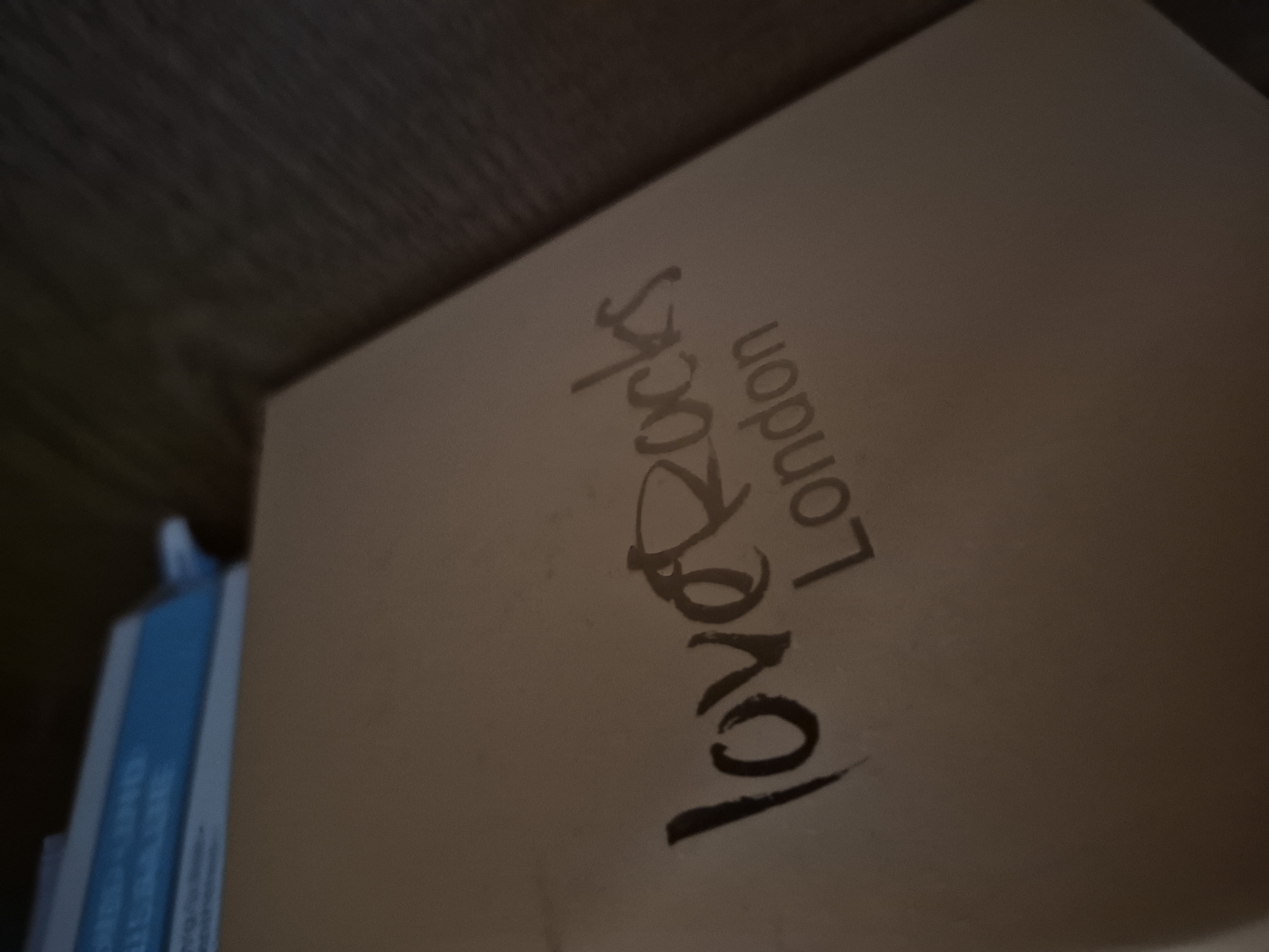 A close-up shot shows the words "love Rocks London" printed on a light brown, textured surface. The text is rendered in two lines; "love Rocks" is written in a stylized, dark font that appears to be brushed or painted, while "London" is printed in a lighter, more subtle sans-serif font below it. The background is dark and out of focus, suggesting a dimly lit environment. The overall impression is that of branding or a design on an object, possibly a box or a book cover.