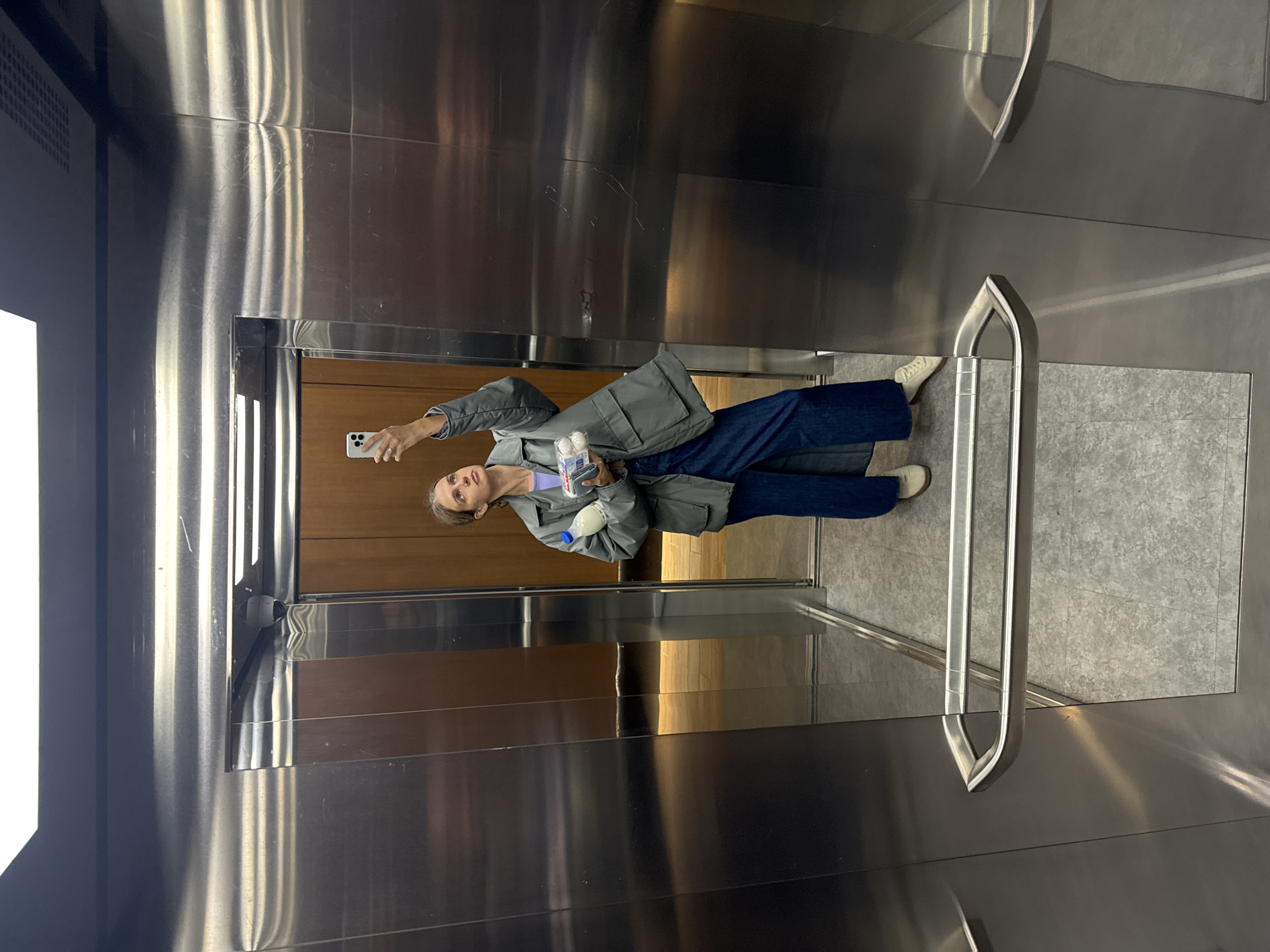 A woman is in an elevator, holding up a phone taking a selfie. She is wearing a gray coat, blue jeans, and white sneakers. She is holding a bottle of water and what appears to be a container of cotton swabs in her other hand. The elevator has a wood-paneled back wall, metal walls and a gray floor. It also has a silver handrail.