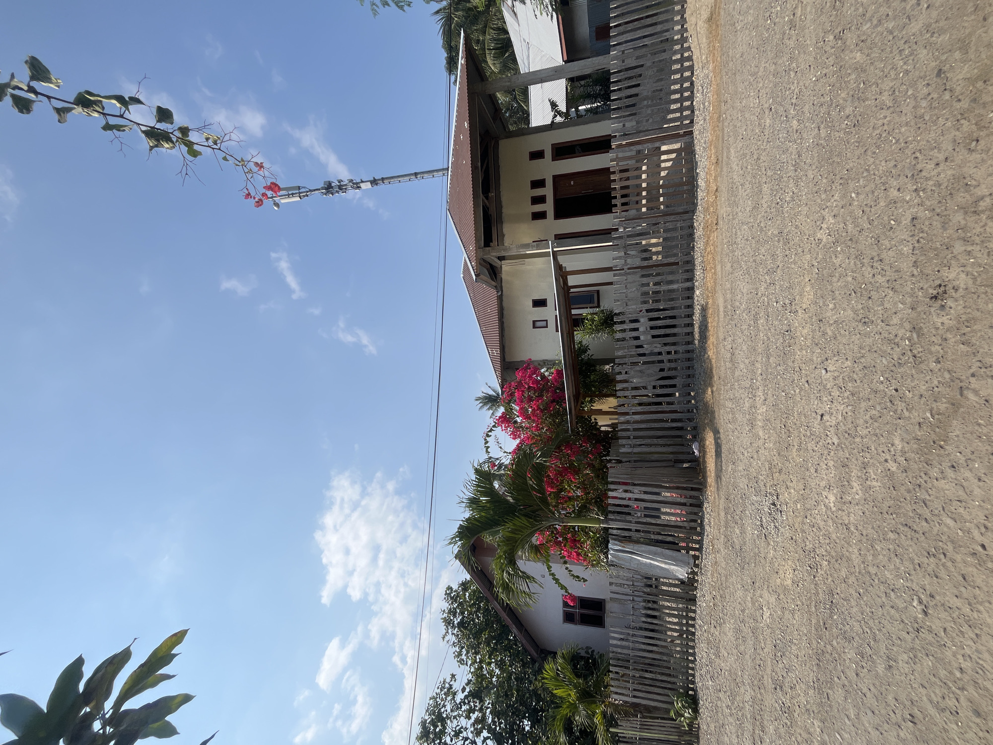 The image shows a view of a residential area with houses, a fence, and a dirt road in Wayipa, Indonesia. In the foreground, a dirt road with loose gravel is visible. Behind the road, a wooden fence with vertical planks encloses the property. Several houses are seen behind the fence, with one house in the center featuring a brown, corrugated metal roof supported by concrete pillars and a covered entrance. This house has several windows and an open doorway. To the left, another house with a similar roofline and a dark-rimmed window is partially visible. Lush green trees and vibrant pink bougainvillea flowers add color to the scene. In the background, a tall communication tower stands against a clear blue sky with scattered white clouds. A branch with green leaves and some red flowers drapes from the top right of the frame, reaching towards the tower. The overall impression is a rural or suburban residential setting.
