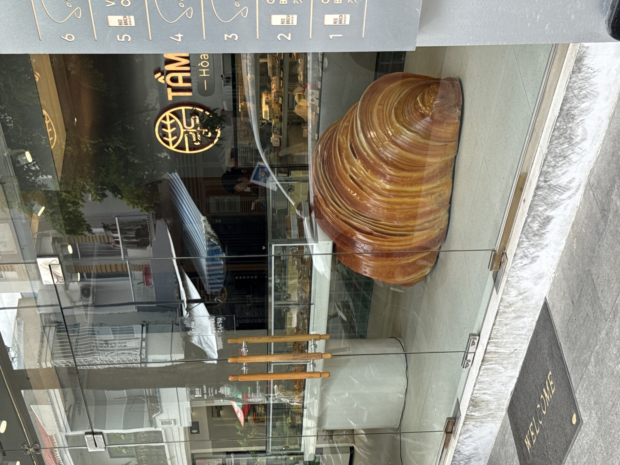A wide shot captures the exterior of a bakery in Nha Trang, Vietnam, as viewed through a glass storefront. The bakery's name, "TÂM HÒA," is visible in gold lettering on a dark background, accompanied by a circular logo featuring a stylized plant or tree. A large, realistic-looking sculpture of a croissant dominates the foreground, placed inside the shop on a tiled floor. Reflections on the glass reveal the urban environment outside, including buildings, trees, and street activity, with a "WELCOME" doormat visible on the sidewalk in front of the entrance. The bakery interior displays glass display cases filled with various baked goods. The lighting is bright, suggesting daytime.