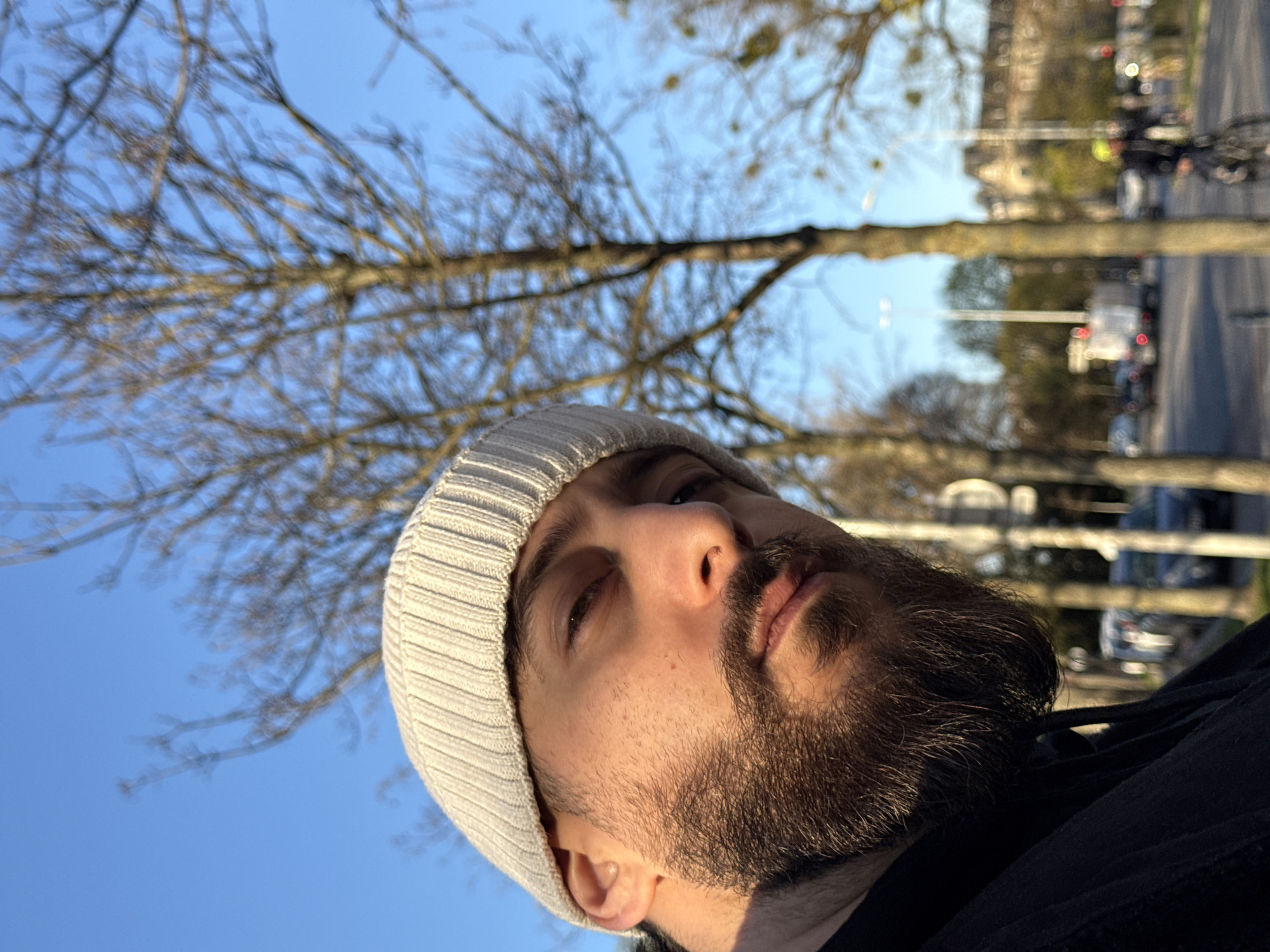 A male individual with a beard and a light beige knitted beanie occupies the left foreground, illuminated by direct sunlight on the right side of his face. He is wearing a dark-colored garment. Behind him, the scene opens to a blurred urban environment under a clear blue sky. Bare deciduous trees, some with mistletoe, line a wide street. Parked cars are visible on the left side of the street, and distant vehicles, including a white truck, are in motion. On the right, a person wearing a bright yellow helmet is riding a bicycle away from the viewer. Multi-story buildings with classic European architecture are discernible in the far distance. The overall setting is consistent with a tree-lined avenue or boulevard in the city of Strasbourg, France, specifically within a district characterized by 19th or early 20th-century architecture, such as the Neustadt or areas near major urban parks.
