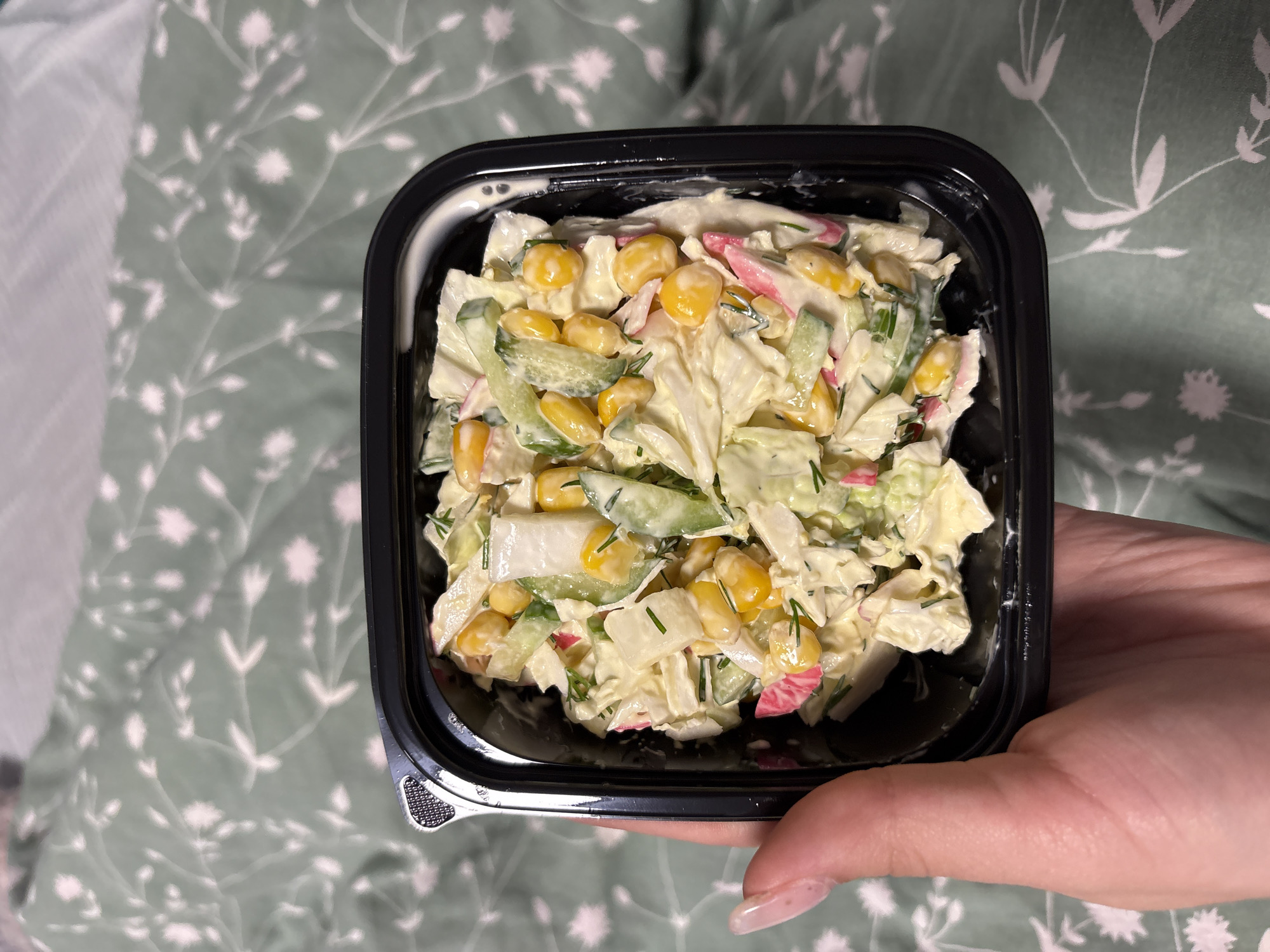 A container of salad is held in a person's hand against a backdrop of a green floral patterned cloth. The salad appears to contain cabbage, corn, cucumber, dill, and imitation crab meat, all coated in a creamy dressing. The container is square, black, and shallow. The person's hand is visible, with a clear nail polish color. The location in the city of Прикамский, Russia is unconfirmed and cannot be assumed.